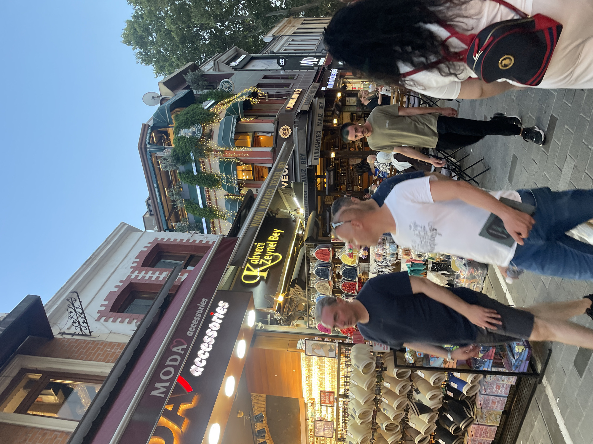 In Kadıköy, Turkey, a street scene captures a bustling pedestrian thoroughfare lined with shops and cafes. The image focuses on the right side of the street, where a group of people are walking. In the foreground, a person with dark curly hair walks away from the camera, carrying a dark backpack with red accents. To their left, a man wearing a light-colored t-shirt and dark pants walks towards the camera. Further to the left, a man in a dark t-shirt and gray shorts is walking, and another man in a white t-shirt and jeans is also moving through the scene. Shops with colorful displays of accessories and food are visible, including a sign for "MODA accessories" and another for "Kahveci Zeynep Bey." An upper floor of a building features outdoor seating for a cafe, adorned with string lights and greenery. The overall atmosphere suggests a vibrant commercial and social area with people engaged in shopping and dining.