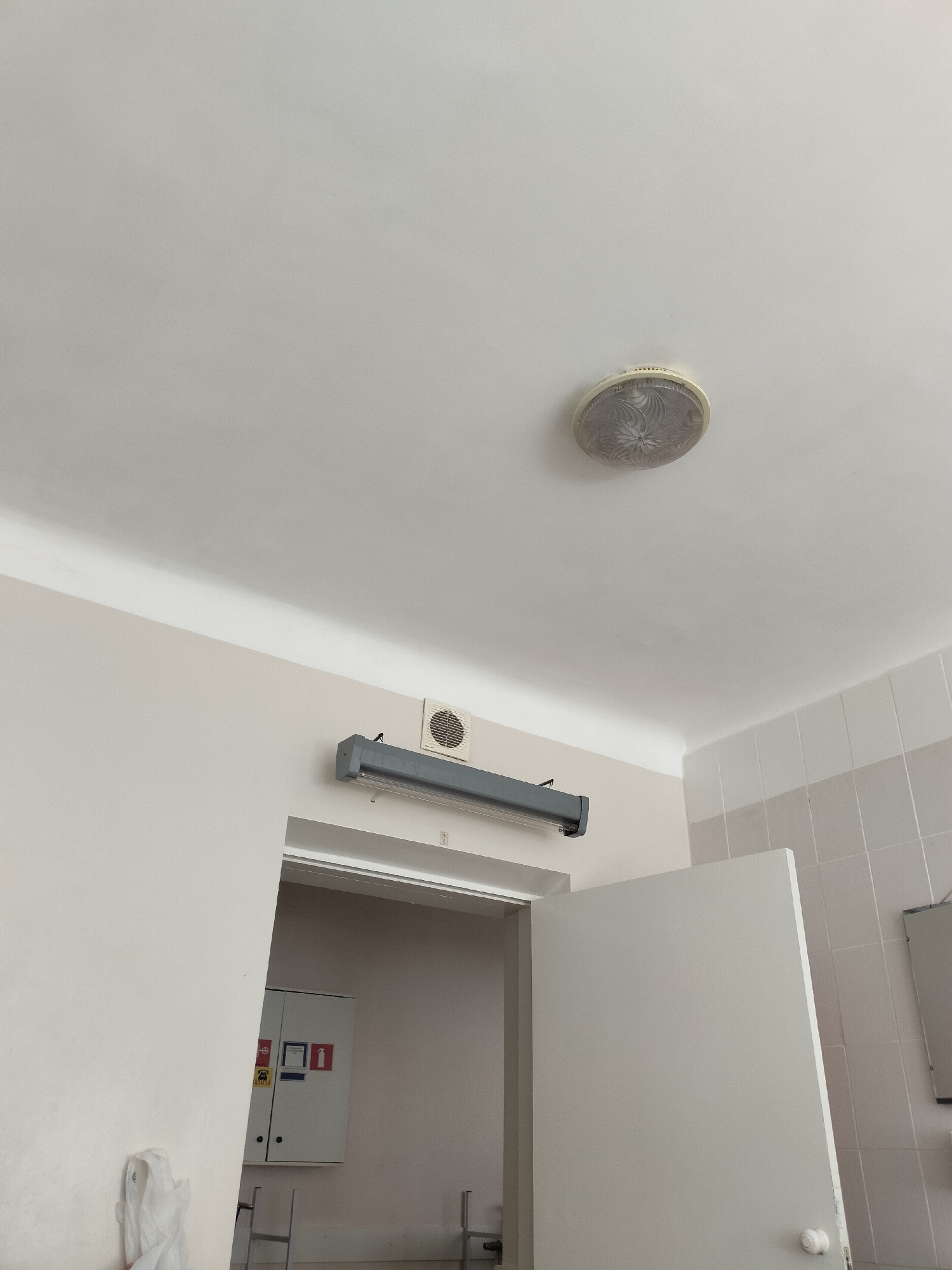 The image depicts an interior view of a room, likely in a public building. The scene includes a white, textured ceiling with a circular light fixture and a long, gray fluorescent light mounted on the wall. Below the light, a ventilation grate is visible. A doorway leads into another room where a white cabinet with various warning signs and labels is mounted on the wall. To the right of the doorway, a white door is ajar, revealing a tiled wall. The overall impression is of a functional, perhaps institutional, space. The specific location in Chernihiv, Ukraine, cannot be determined from the visual information provided.