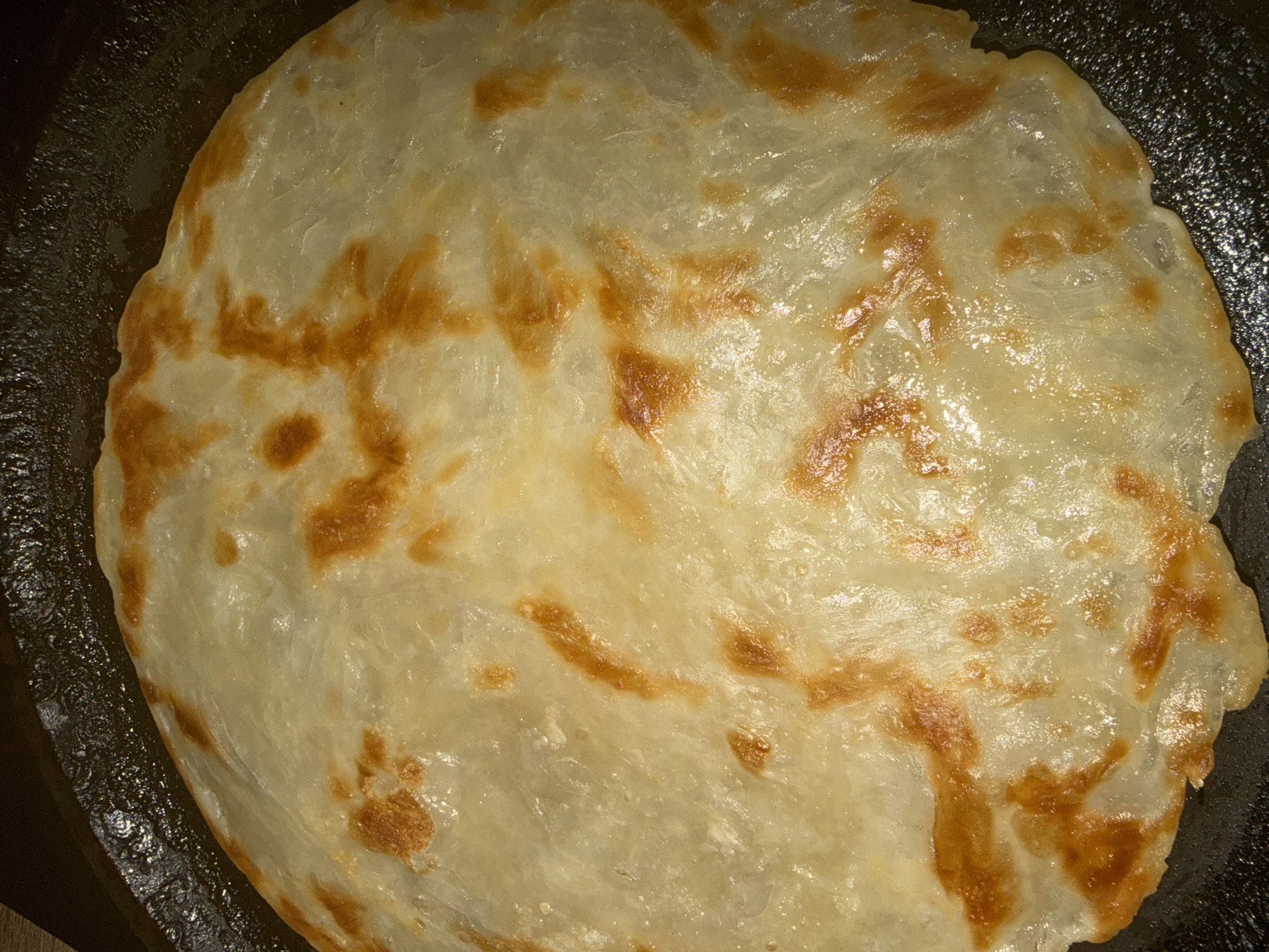 A flatbread-like food is cooking in a dark, possibly seasoned pan. The flatbread is round with a slightly irregular edge and appears to be made of layers, possibly dough or pastry. It has a light, translucent appearance with scattered patches of golden-brown coloring suggesting areas of higher heat contact with the pan. The surface appears slightly oily or moist. The background is minimal, showing only the edges of the pan. There is no indication of the location being Chaikovsky, Russia or any other specific location.