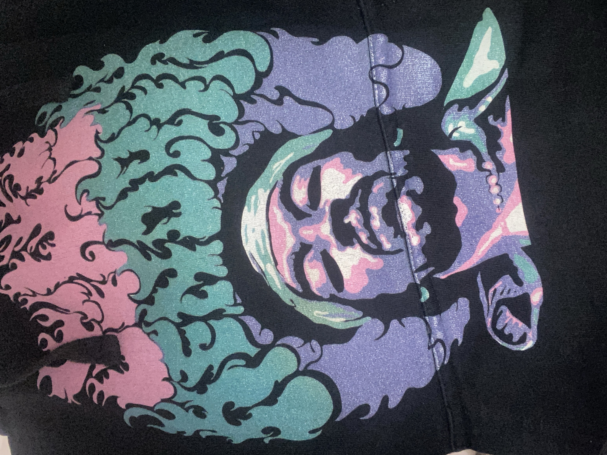 A close-up image displays a vibrant, multi-colored graphic design printed on black fabric, likely part of a garment. The central element of the design is a highly stylized, abstract face or skull presented in profile, facing towards the right. This face is rendered in shades of lavender, pastel pink, and white, with subtle light green accents, and features prominent eye sockets, a defined nose area, and an open mouth. Surrounding the face are dynamic, swirling patterns reminiscent of clouds, flames, or intricate natural forms. These patterns are depicted in soft pink and a shimmering teal or mint green, with distinct black outlines that enhance their complex shapes. A visible vertical seam or fold runs down the center of the graphic, passing through the stylized face. The print itself exhibits a slightly textured, granular quality, characteristic of screen printing. The overall aesthetic is abstract and fantastical, utilizing a palette of bright, contrasting pastel colors against the deep black background. No discernible activity is taking place, as it is a static image of a design. There is no visible text.