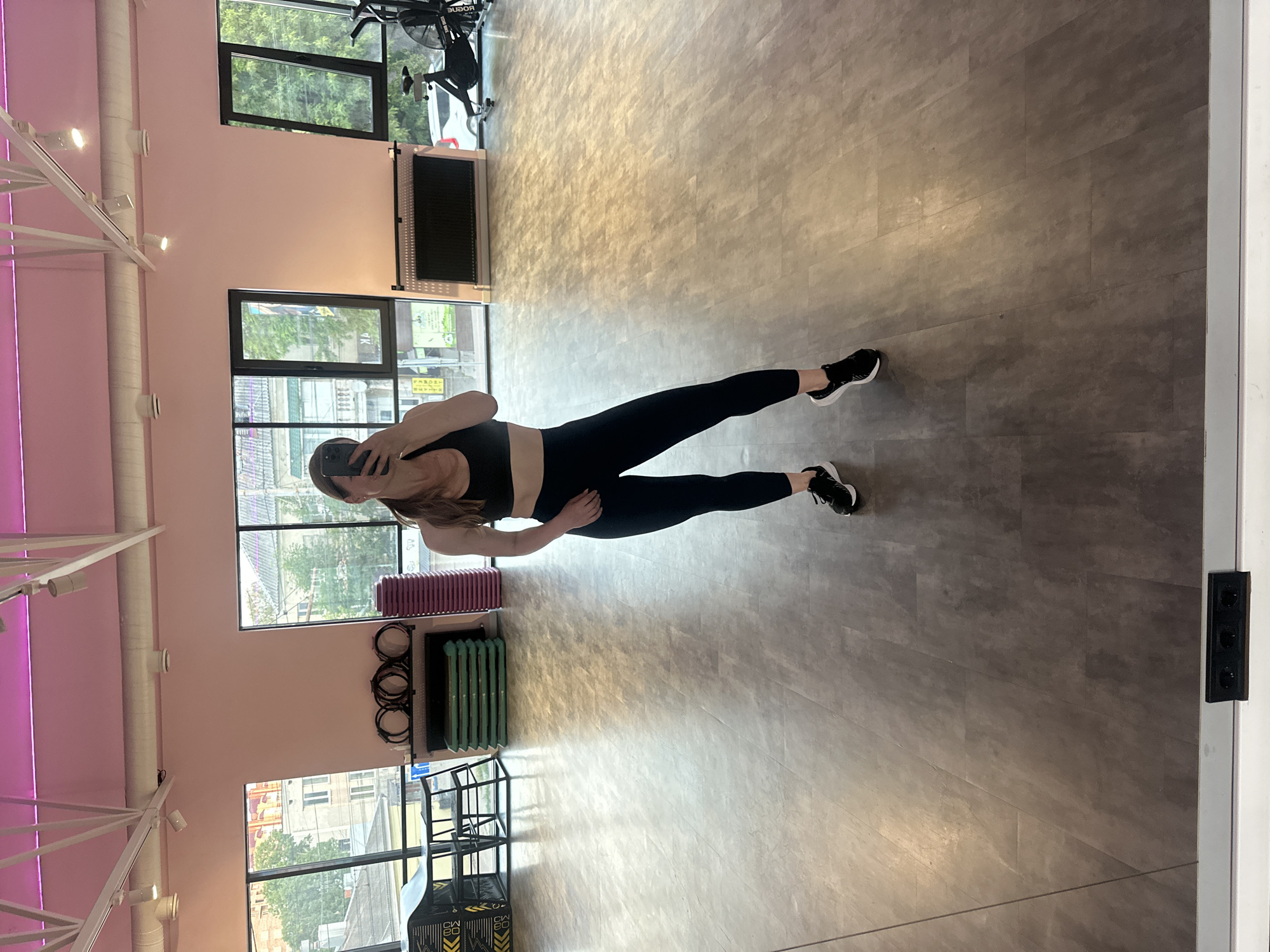 A full-body, mirror selfie of a woman in athletic wear is captured in a gym. She is wearing a black sports bra and black leggings, with black and white athletic shoes. The woman is standing on a grey, wood-patterned floor. Behind her, visible in the reflection of a large mirror, are gym equipment, including a stationary bike, step platforms, and what appear to be weighted vests or similar workout gear. Large windows offer a view of buildings and trees, suggesting an urban setting. The gym features pink painted walls and a pink ceiling with white industrial lighting fixtures. A heating unit is mounted on the wall. The overall impression is of a modern fitness studio. The location is identified as a gym in Ternopil, Ukraine, based on context provided.
