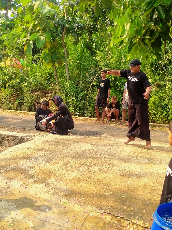 The image depicts an outdoor scene in Gebyog, Indonesia, under bright daylight, featuring six individuals engaged in a group activity. In the foreground, a barefoot male, wearing a black t-shirt with white text and a black baseball cap, is captured mid-air, jumping over a rope. His right arm is extended forward. To his left, another barefoot young male, clad in a black t-shirt and shorts, holds one end of the jump rope, standing on a paved road. Further left on the same paved road, a person in a black long-sleeved garment and a black head covering is crouched low, facing a seated male next to them. Behind the rope-holding individual, another young male squats barefoot. A sixth person is partially visible behind the jumping male. The background consists of dense, vibrant green foliage and various trees. The immediate foreground is a light-colored concrete surface with scattered small pinkish debris, separated from the darker paved road by a low concrete retaining wall. A partial blue plastic barrel is visible in the lower right foreground.