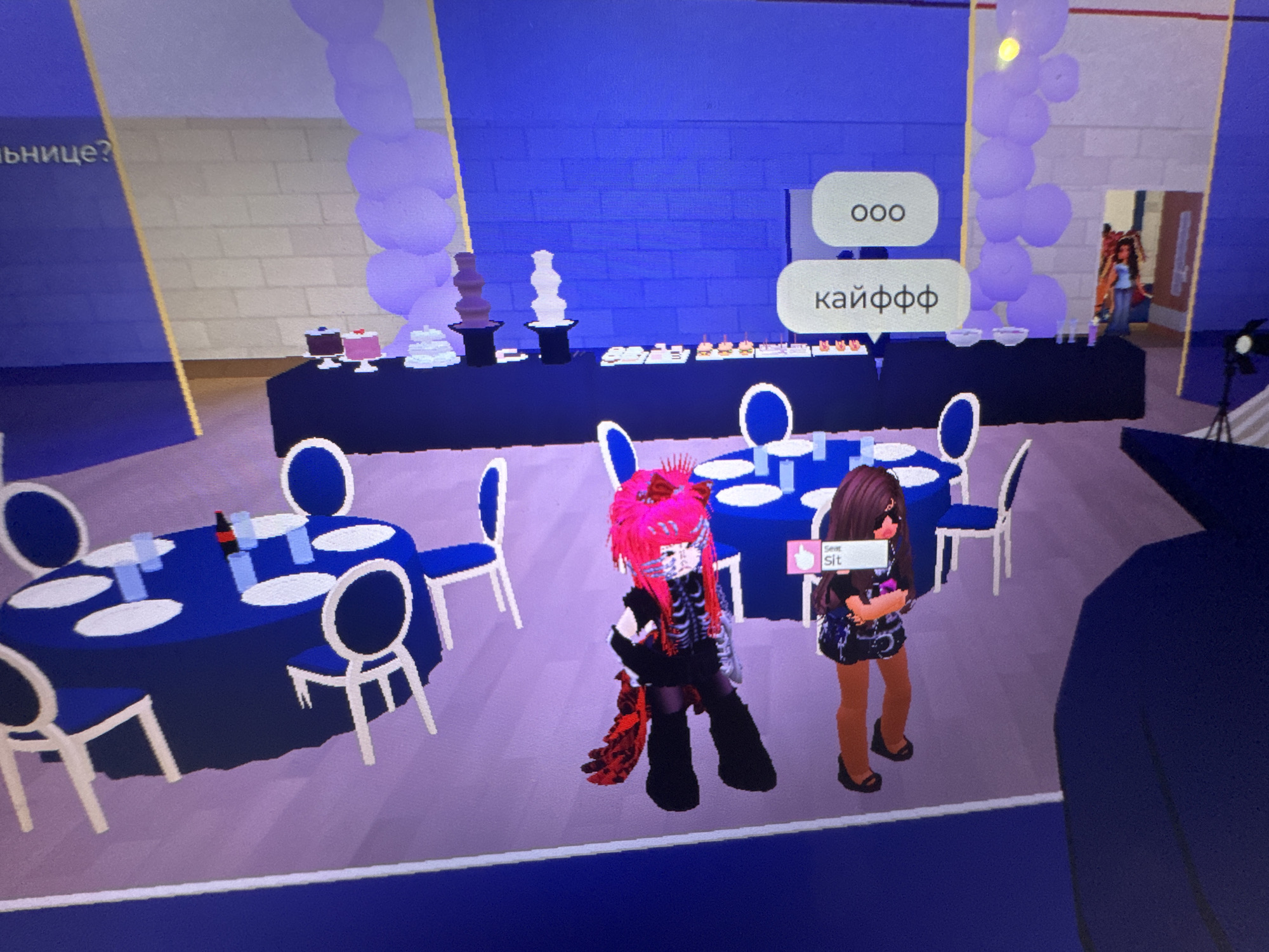 An indoor scene, likely from a video game like Roblox, depicts an event space set up for a party or reception. The room features a prominent buffet area and multiple dining tables.
The central buffet table is covered with a dark tablecloth and laden with various desserts, including two small cakes (one chocolate, one pink), a tiered white dessert stand, and two multi-tiered chocolate fountains (one dark chocolate, one white chocolate), alongside other small pastries. Behind the buffet, the wall is a textured blue, flanked by tall columns of light purple balloons.
To the left, two round dining tables are covered with royal blue tablecloths and set with white plates and blue glasses. White chairs with blue seat cushions are arranged around these tables. The floor is a light purple, complementing the overall blue and purple color scheme.
Two female avatars are in the foreground. The character on the left has vibrant pink hair, a black outfit with a white skeleton design, and red accents, including a feathery tail-like attachment. The character on the right has long brown hair, dark sunglasses, a patterned dark mini-dress, and black wedge shoes.
Speech bubbles above the pink-haired character display Russian text: "ООО" (an interjection like "oh" or "wow") and "кайффф" (slang meaning "awesome" or "delight"). Above the brown-haired character, a prompt reads "Seat" and "Sit" next to a thumbs-up icon, suggesting an interaction.
In the far background, a doorway reveals a glimpse of another character. A camera on a tripod is also visible on the far right. The lighting is bright and even, indicating an indoor, possibly artificially lit, setting. Partially visible text on a blue panel on the far left reads "льнице?".
