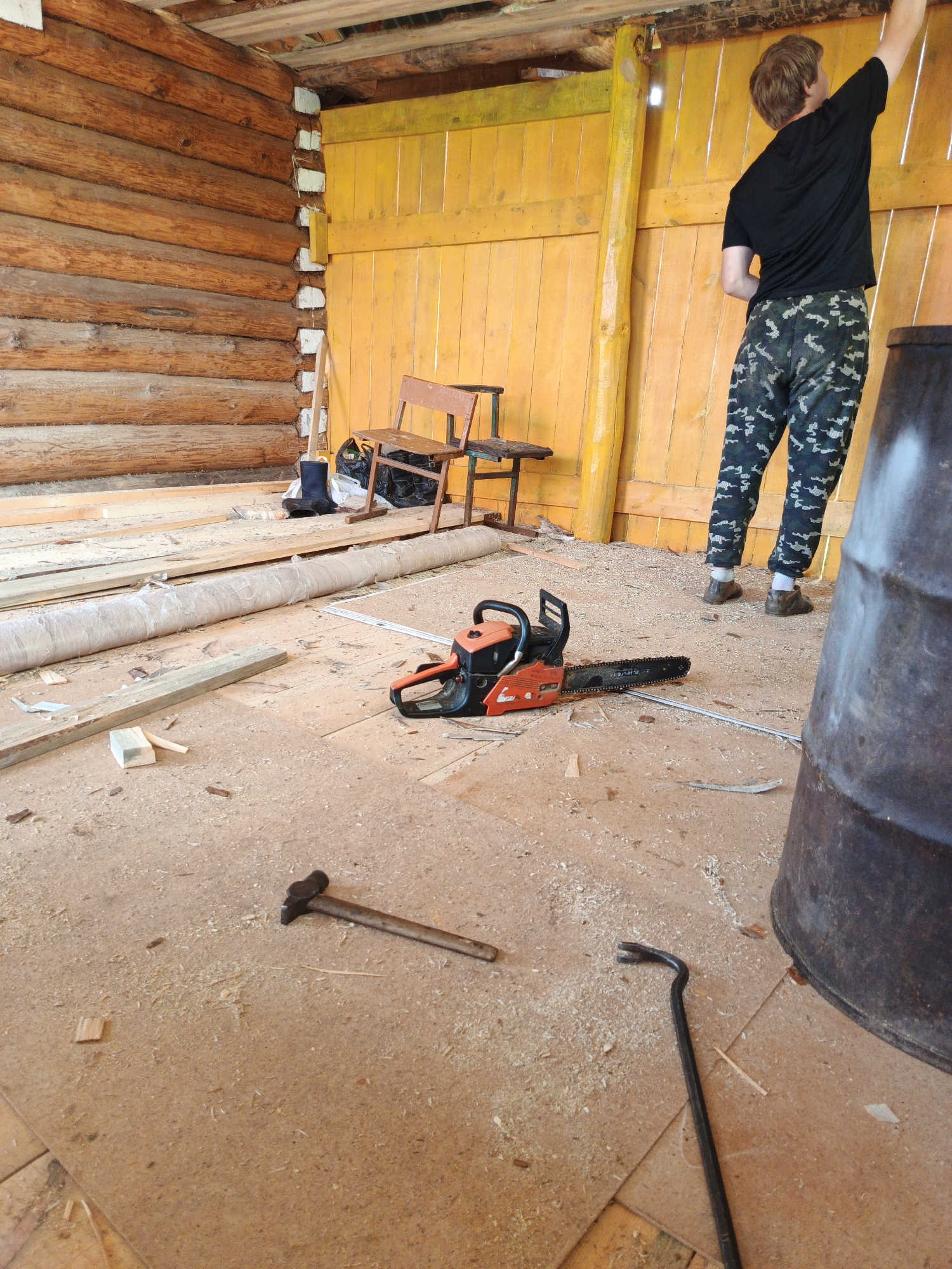 The image depicts the interior of a wooden structure, possibly a barn or unfinished building, located in Islambaevo, Russia. A young man, wearing a black t-shirt and camouflage pants, is standing and reaching upwards, seemingly engaged in work on the ceiling. In the foreground, a bright orange and black chainsaw rests on a floor covered in wood shavings and scattered planks. Other tools, including a hammer and a crowbar, are also visible on the floor. A large, dark barrel is situated on the right side of the frame. The walls are constructed of rough-hewn logs, and the background features yellow wooden paneling. The overall scene suggests ongoing construction or renovation work.