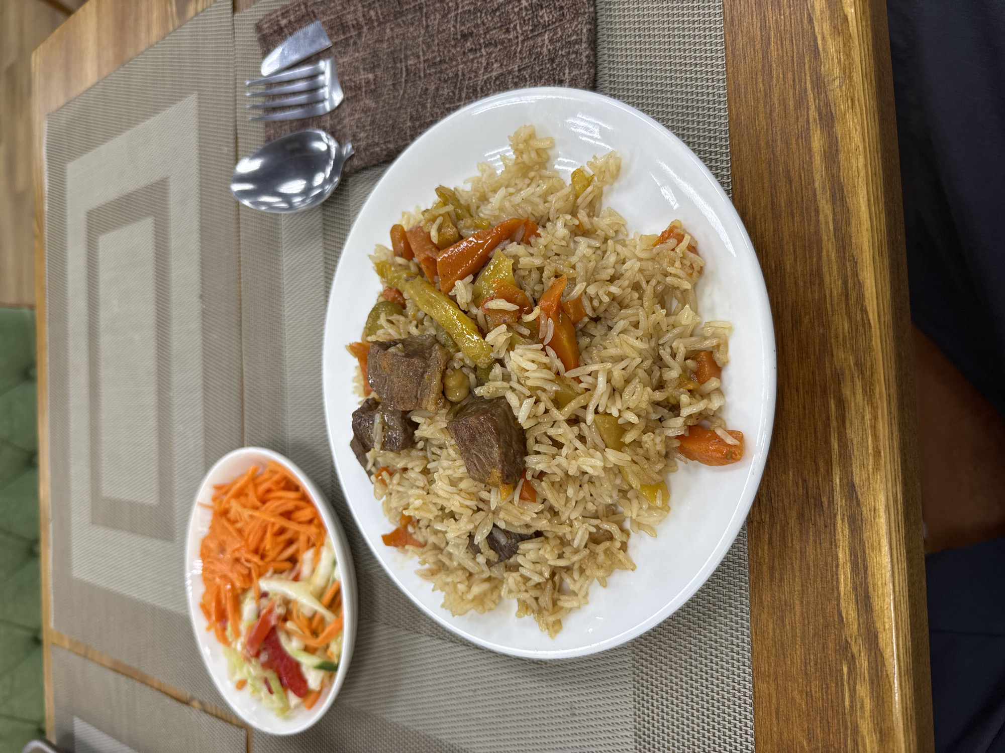 A top-down shot shows a white plate filled with pilaf, a rice dish often made with meat and vegetables. The pilaf contains rice, chunks of meat, and pieces of carrots and other vegetables. To the left of the plate is a small white bowl with a side salad, primarily composed of shredded carrots and some other chopped vegetables. Cutlery, a fork and a spoon, are placed on a brown placemat to the upper left of the plate. The scene is set on a wooden table, with a textured placemat underneath the food. The lighting suggests it could be daytime. No visible text is present. The setting appears to be an indoor dining area, possibly a restaurant, given the presentation of the food and table arrangement. The overall mood is one of a prepared meal, ready to be eaten. The context provided indicates this meal is likely in Moscow, Russia.
