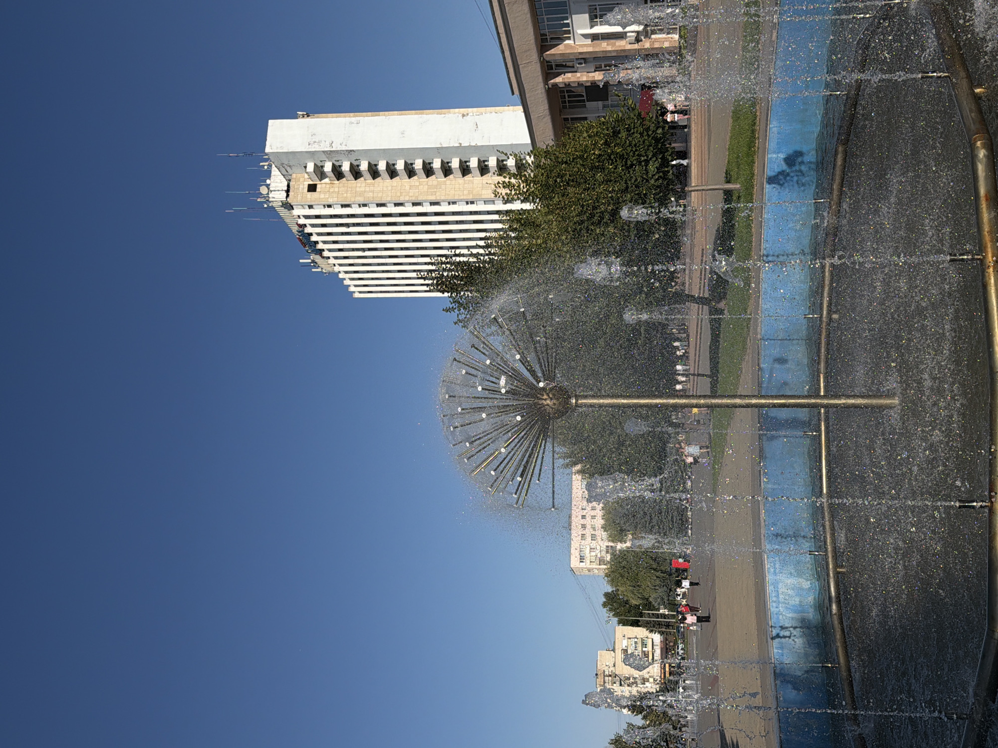 The image depicts a public space in Volzhsky, Russia, featuring a modern-style fountain in the foreground. The fountain has a metallic, spherical head with numerous thin rods radiating outwards, from which water sprays in a fine mist, creating a dandelion-like effect. The fountain basin is circular with blue-painted walls and is filled with water reflecting the spray. Behind the fountain, several trees with lush green foliage are visible, partially obscuring the view of buildings.
In the background, there is a prominent, tall, multi-story building with a light-colored facade, characterized by vertical white and beige stripes. The building has numerous windows and balconies and is topped with several antennas. To the right of this building, another, lower building with a light brown facade and white columns is partially visible.
A paved area, possibly a plaza or sidewalk, stretches behind the fountain and trees. Several people are walking in this area, their figures small in the distance, suggesting a public park or urban square. The sky above is a clear, vibrant blue, indicating fair weather. The overall scene captures a bright, sunny day in an urban environment, likely a public gathering space with recreational features.