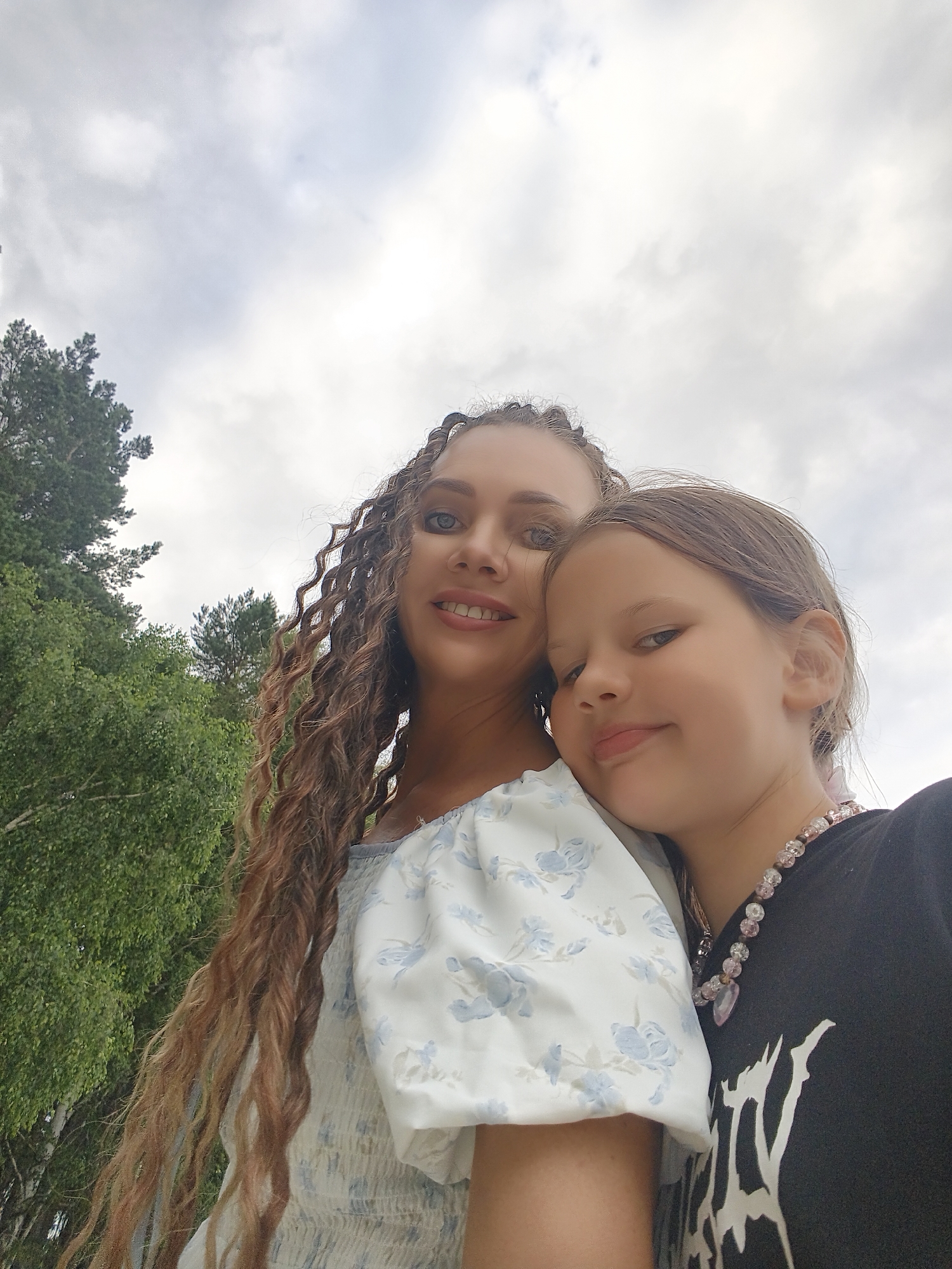 This is a close-up selfie of two females, presumably mother and daughter, taken outdoors under a cloudy sky. The younger female on the right rests her head on the shoulder of the older female on the left. The older female has long, curly brown hair and is wearing a white dress with a blue floral pattern. The younger female has brown hair and is wearing a black t-shirt with a white graphic and a beaded necklace. Green trees are visible in the background, suggesting a natural setting, possibly a park or residential area. The image is taken from a low angle, looking up towards the sky. The overall impression is a candid, affectionate moment shared between the two individuals.