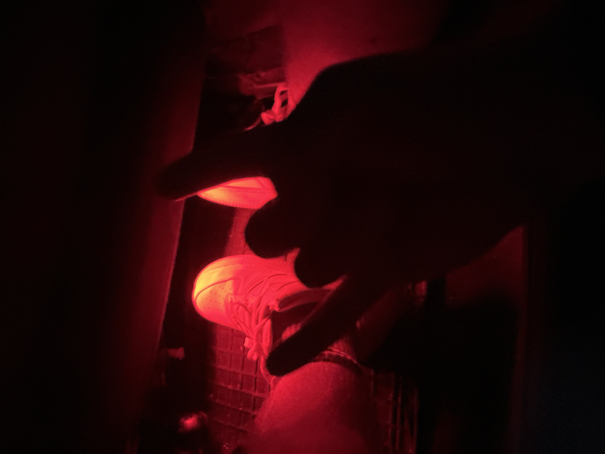 The image captures a dimly lit interior, bathed in a strong red glow. In the foreground, a hand with fingers extended forms a silhouette against the light. Partially visible beneath the hand is a person's feet, shod in light-colored sneakers, resting on a dark, textured surface, possibly a car's footwell. The red lighting creates an atmospheric and somewhat abstract scene, obscuring fine details of the surroundings. The location in Zlatograd, Bulgaria, is not visually discernible from the image itself.