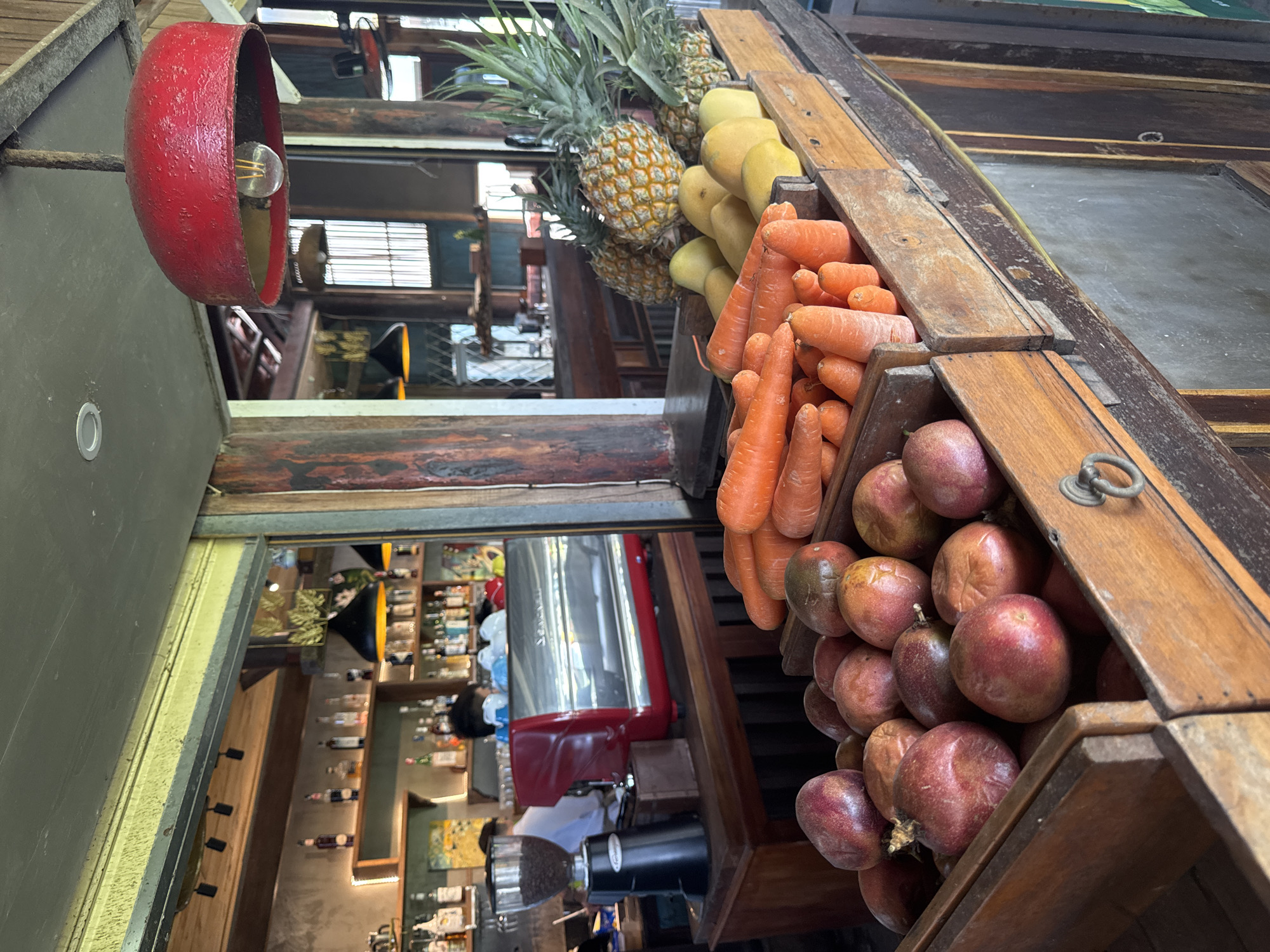 A high-angle interior shot shows a display of fruits and vegetables, including pineapples, mangoes, carrots, and passion fruits, presented in wooden crates. A red, bowl-shaped lamp hangs above. The background reveals a bar or cafe setting with shelves stocked with liquor bottles, a red espresso machine, and a coffee grinder. The location is Đà Nẵng, Vietnam.
