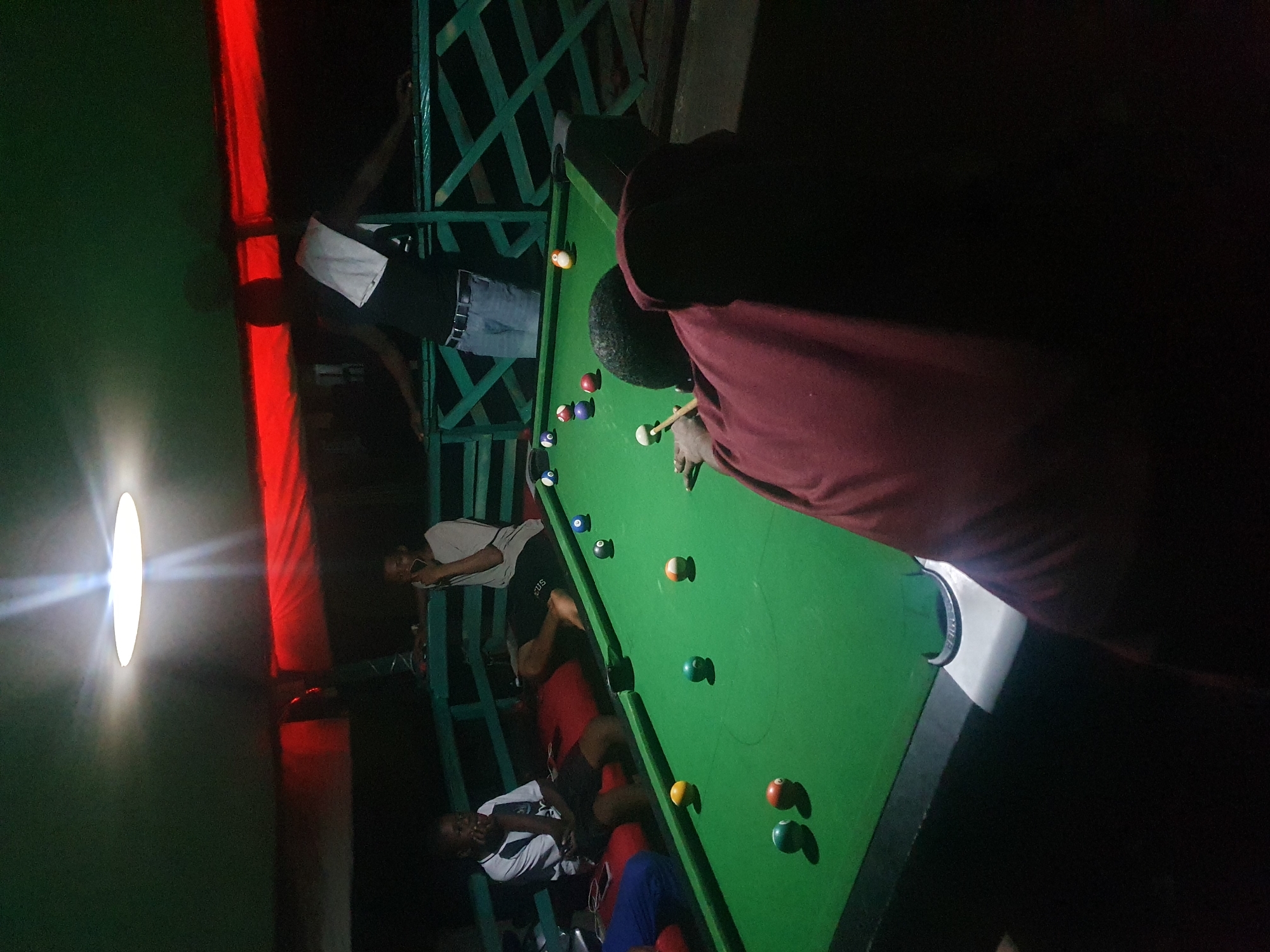 The image depicts a group of young men playing pool at a dimly lit establishment, possibly a bar or a recreational center, located in Lokoja, Nigeria. The scene is set at night, with a bright overhead light illuminating the pool table and casting long shadows. The pool table itself is covered in green felt, with several billiard balls scattered across its surface. One person, wearing a maroon garment, is actively engaged in playing, holding a pool cue and aiming at a white ball. Other individuals are visible in the background, seated and observing the game. The environment appears to be an open-air or semi-enclosed space, indicated by the presence of a railing and what seems to be a canopy or roof overhead. The red fabric draped in the background suggests decorative elements or temporary partitioning. The overall atmosphere is casual and social, with the focus on the game of pool.
