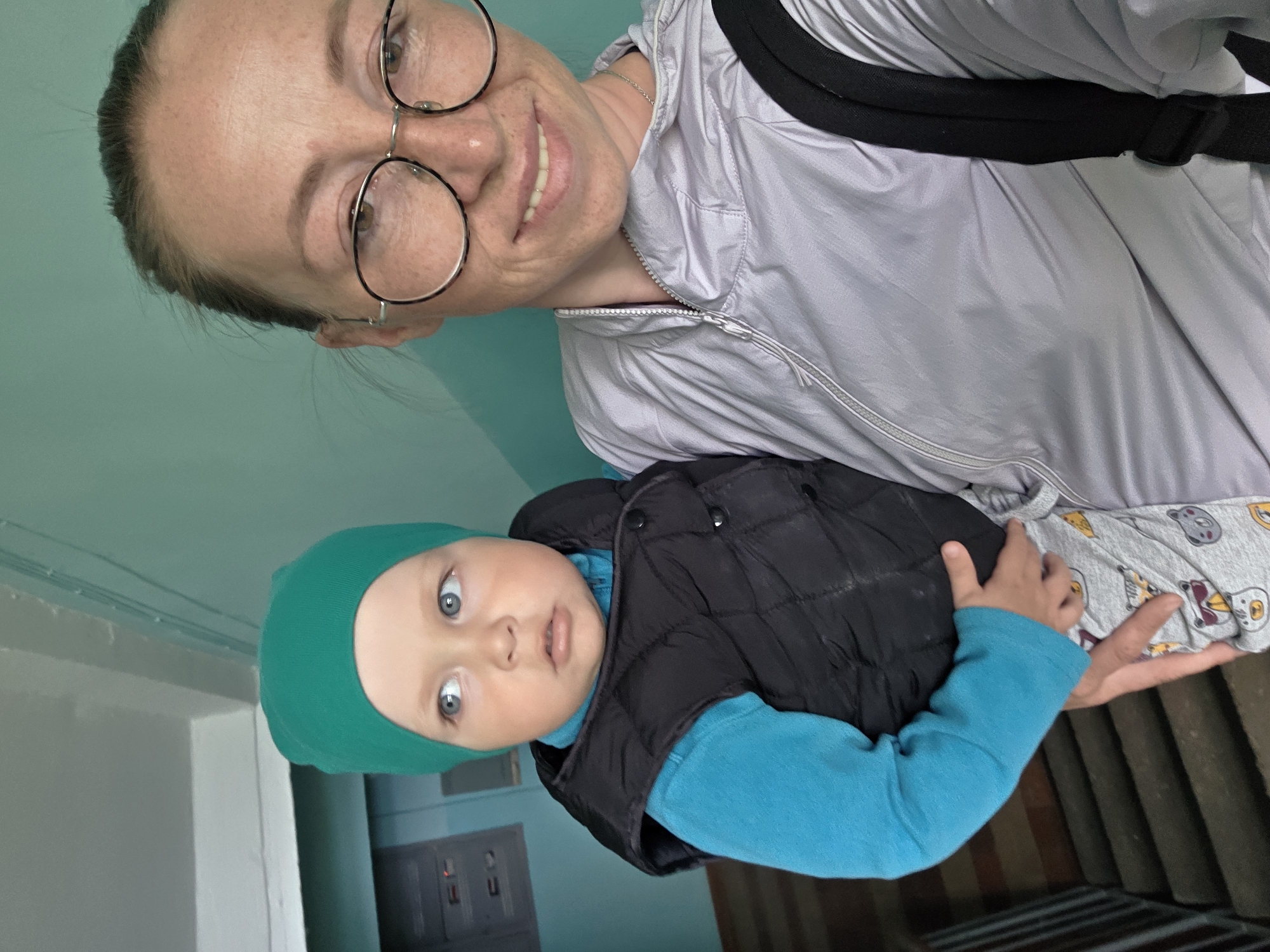 A woman with round glasses and a light gray jacket is holding a baby wearing a green hat, a blue long-sleeved shirt, and a black vest. The location appears to be indoors, with walls painted in shades of green and white. A staircase is visible in the background. The specific location is Chaikovsky, Russia.