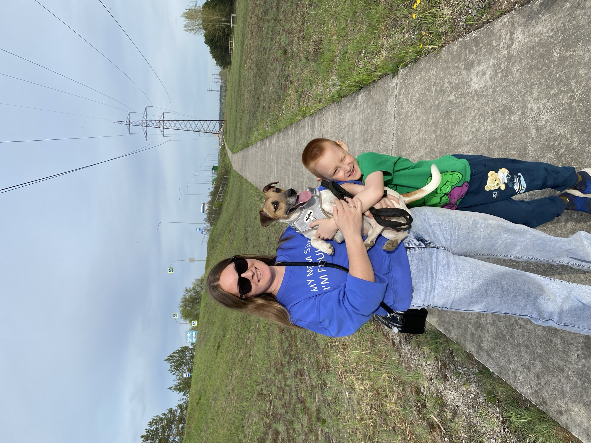 A woman, a young boy, and a dog are standing on a concrete path in what appears to be a public space in Noviy, Russia.
The woman is holding the dog. She is wearing sunglasses, a blue sweater with white lettering, and light-colored jeans. She has a black purse hanging from her shoulder. The dog is wearing a gray harness.
The young boy is wearing a green shirt and dark blue pants with cartoon characters. He is touching the dog and smiling.
The background includes grassy areas on either side of the path, a row of street lights and road signs, and power lines supported by metal towers. The sky is overcast.