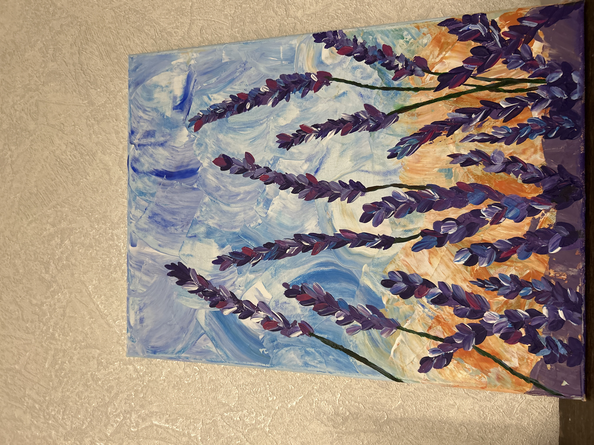 The image displays a vibrant, textured painting of lavender flowers on a canvas, leaning against a textured, light-colored wall. The painting features numerous stalks of lavender, depicted with prominent brushstrokes and impasto, primarily in various shades of purple, blue, and reddish-purple. The stalks themselves are painted in dark green.
The background of the painting is abstract and divided: the left side consists of swirling blues and whites, evoking a sky or water, while the right side transitions into warm hues of orange, yellow, and some darker purples, suggesting a sunset or a field at dusk. The overall style of the painting appears to be impressionistic or abstract, with visible paint textures adding depth and character to both the flowers and the background.
No specific activity is taking place other than the display of the artwork. There are no people visible. The setting is indoors, indicated by the wall texture. No time of day or specific weather conditions are discernible from the scene. There is no visible text in the image.