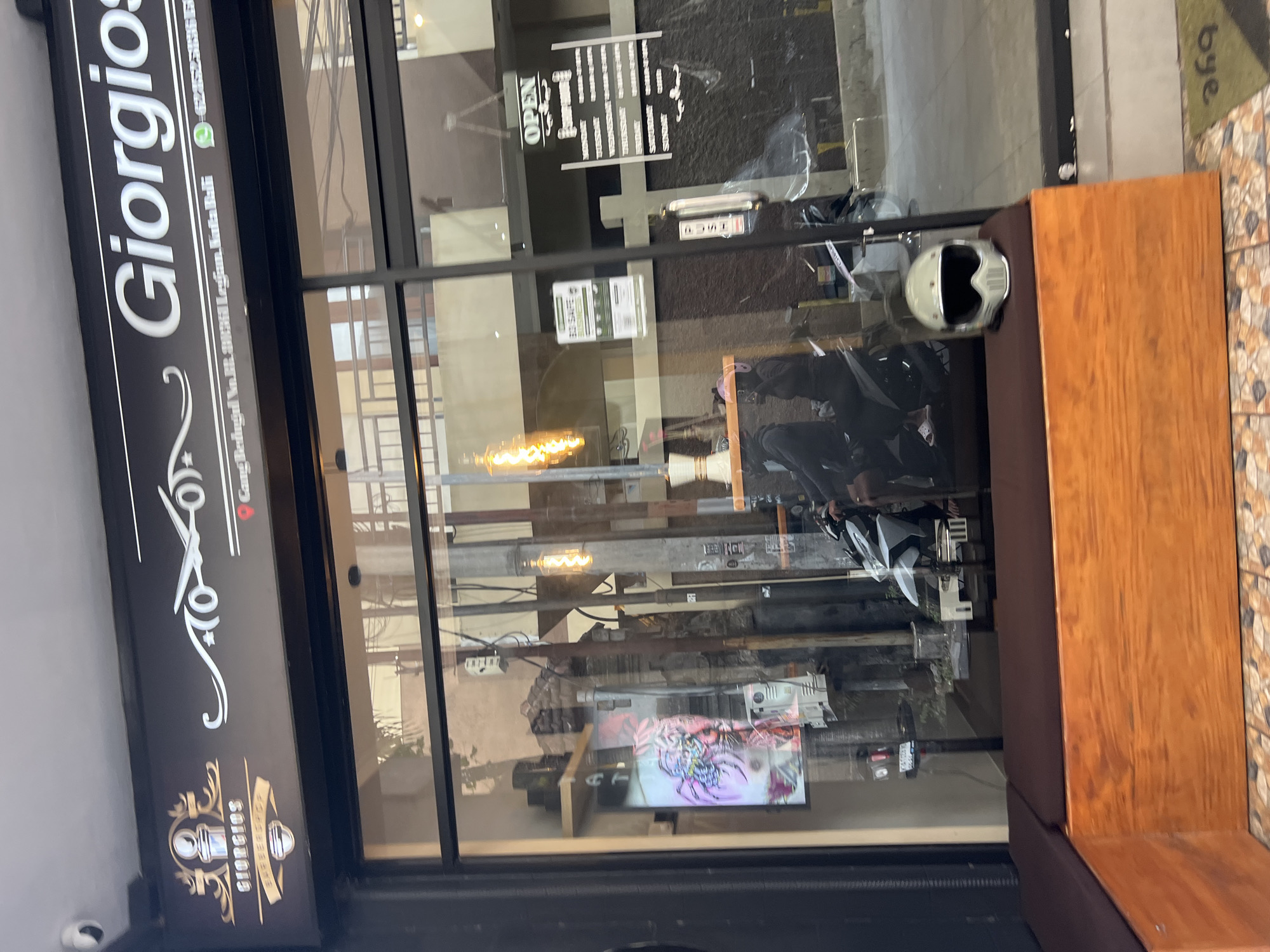 The image shows the exterior of a barbershop called "Giorgios Barbershop" in Kuta, Indonesia. The barbershop's sign is prominently displayed above the entrance, featuring the name in white lettering against a black background, with decorative elements and a logo. Below the name, the address "Gang Bedugul No.88, 80361 Legian, Kuta, Bali" is visible.
The entrance appears to be a glass door, with reflections of the interior and the street visible on its surface. Inside, a digital display shows a colorful graphic. There are various items visible through the glass, including shelves with boxes, electrical wires, and light fixtures. A motorcycle is parked inside, and two people are seated on it. Another motorcycle is partially visible in the background, and a person is standing next to it. A white helmet rests on a wooden bench outside the shop. A sign with "OPEN" is hanging on the glass. Another sign with a QR code and text related to "BE SAFE BUSINESS" is also visible. The flooring outside the shop is tiled.