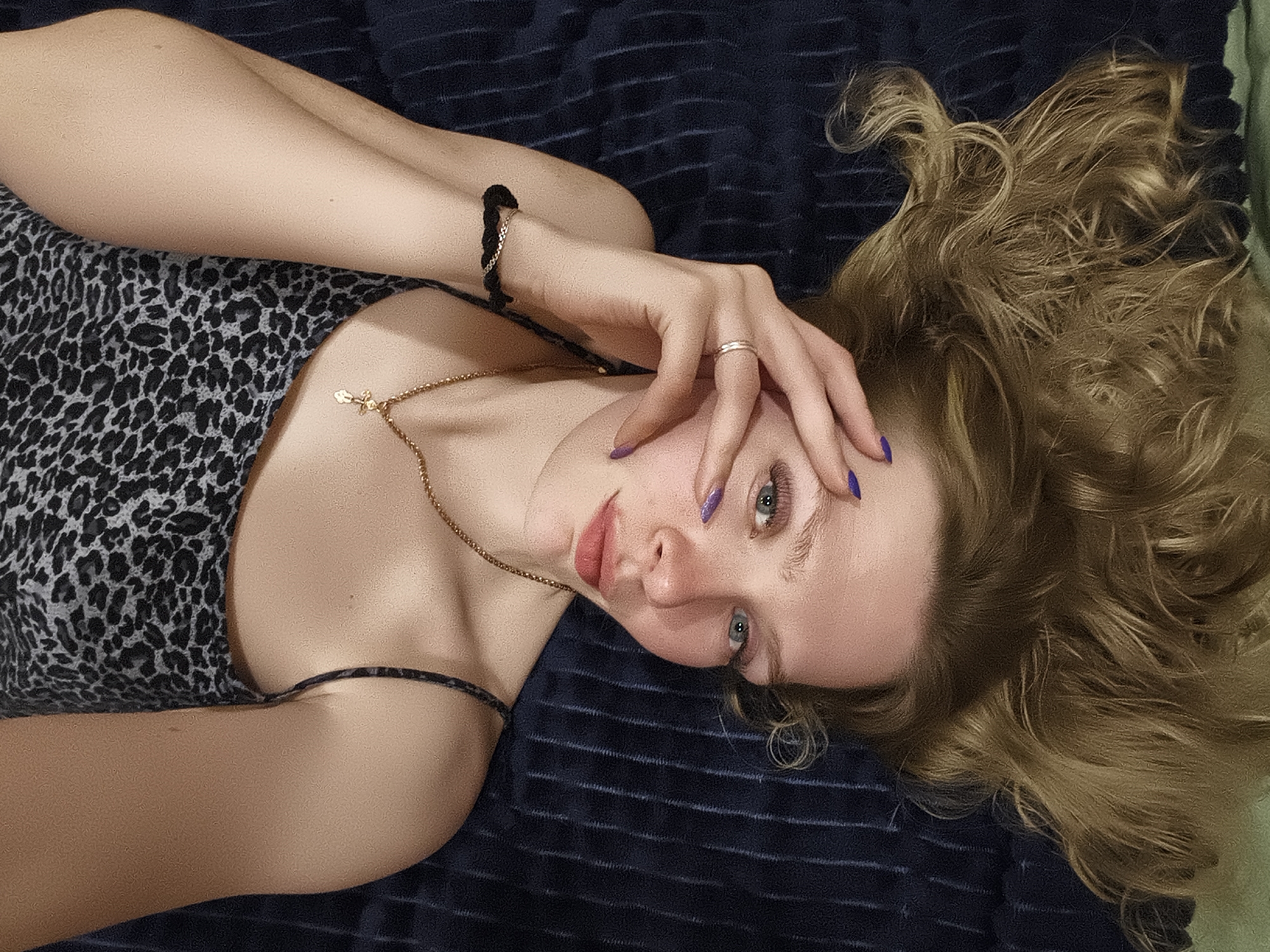 A young woman with fair skin and blonde, curly hair is lying on a dark blue striped blanket. She is wearing a grey and black spotted top with thin straps and a gold necklace with a small cross pendant. Her left hand is resting on her forehead, and she has a ring on her finger and a black bracelet on her left wrist. Her nails are painted a blueish purple. The location is unknown, and there is no evidence to suggest it is in Русский Вожой, Russia.