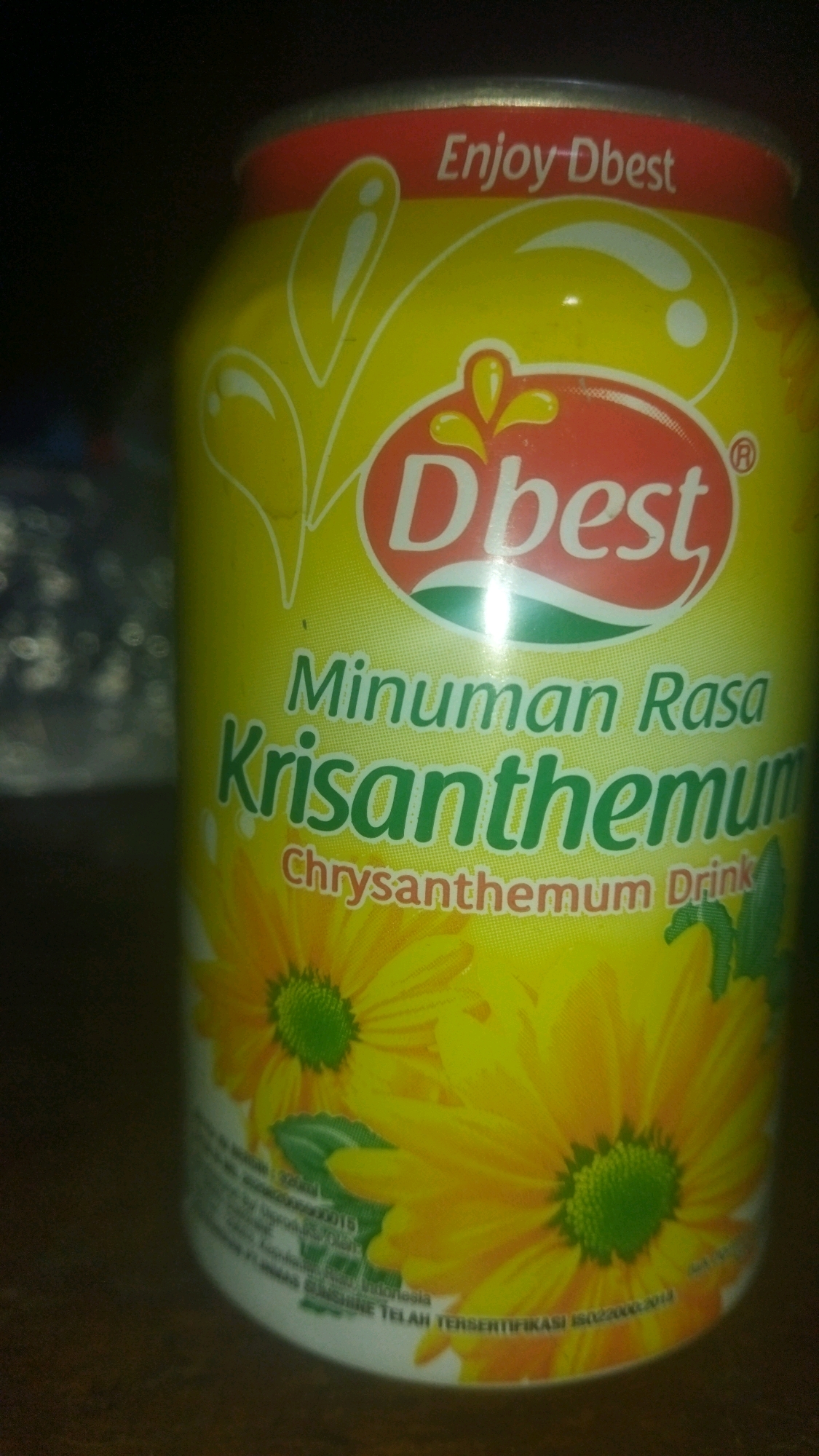 The image shows a close-up of a yellow aluminum can with red and white branding. The can has an illustration of yellow flowers and green leaves. Text on the can reads "Enjoy Dbest" and "Minuman Rasa Krisantemum" which translates to "Chrysanthemum Drink". The can is sitting on a dark wooden surface. The background is dark and out of focus. There is no indication of the location in Tarai Bangun, Indonesia within the image.