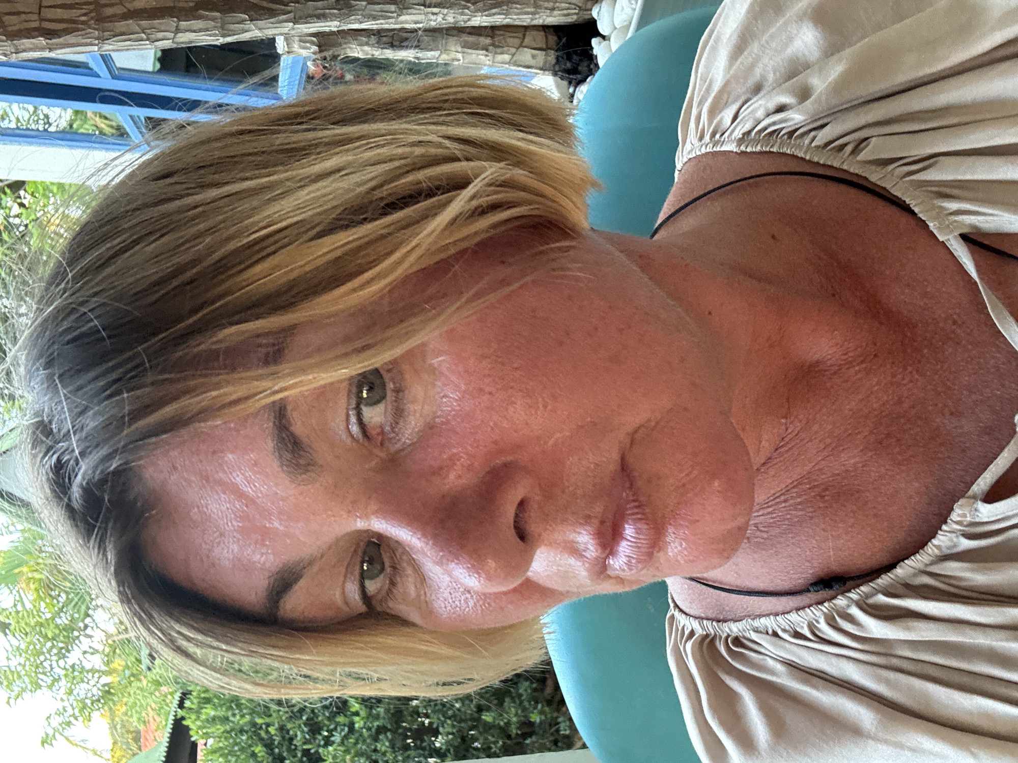 A close-up, vertically oriented shot shows a fair-skinned woman with short, blonde hair. The skin on her face and neck appears flushed, possibly sun-affected. She has green eyes. She wears a beige, silky top gathered at the neckline with a thin, dark cord that seems to act as a necklace. The background includes greenery, possibly trees or shrubs, and glimpses of structures, indicating an outdoor or semi-outdoor setting. Based on the prompt information, this location is in Baan Bo Phut, Thailand.