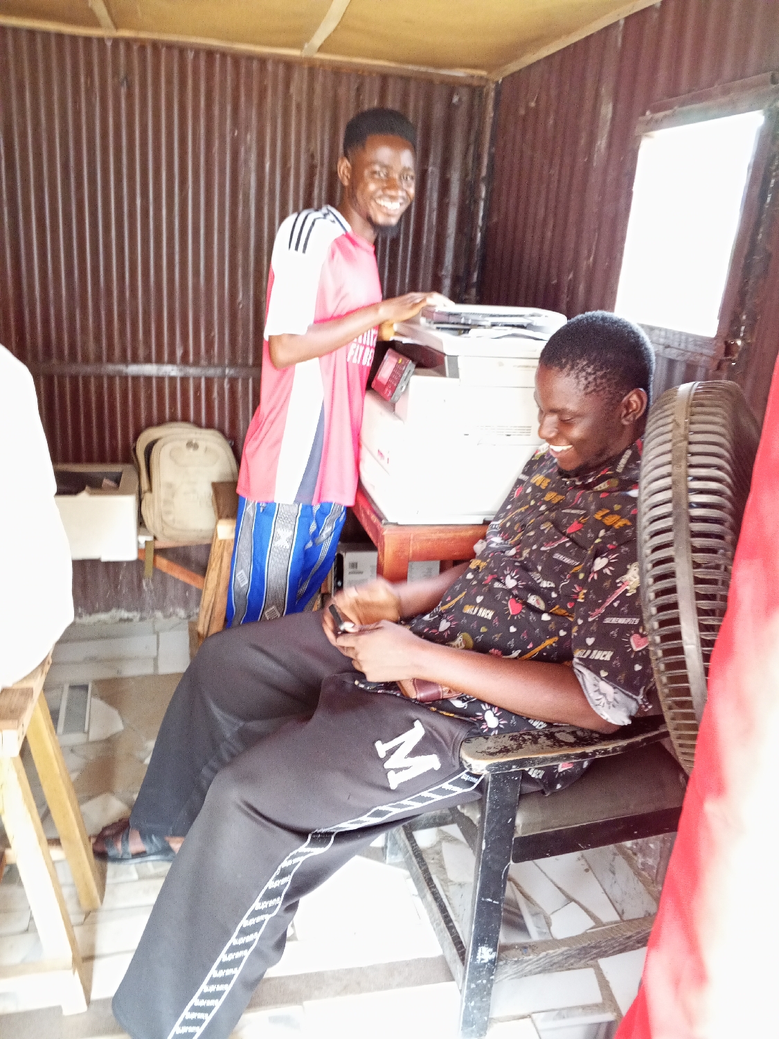 A vertical shot captures two young men inside a small, functional room, likely a shop or office in Zango-ndaji, Nigeria. The interior walls are constructed from corrugated metal sheets, and bright daylight streams in through a small, rectangular window on the right side.
In the center-left, a man stands behind a large white multifunction printer or copier machine, with his hands positioned near its top surface. He wears a red and white short-sleeved t-shirt with black stripes on the shoulders, featuring partially visible text "FLY BE...", and blue and white patterned shorts. He sports a short, dark beard and a wide, friendly smile, looking towards the viewer. A light-colored backpack is visible on a stool behind him.
To the right, another man is seated in a dark metal chair, looking down at his black smartphone. He is dressed in a dark, patterned short-sleeved shirt adorned with small designs, including text like "WILD ROCK", and black sweatpants. The sweatpants have a prominent white "M" on the left thigh and a white side stripe with repeating text, possibly "supreme". He also smiles broadly, indicating a cheerful and relaxed mood. A dark-colored standing electric fan is partially visible to his right, suggesting a warm setting.
Both men appear happy and at ease. The scene depicts a casual, friendly atmosphere in what seems to be a local business offering printing or copying services. The bright lighting suggests it is daytime.