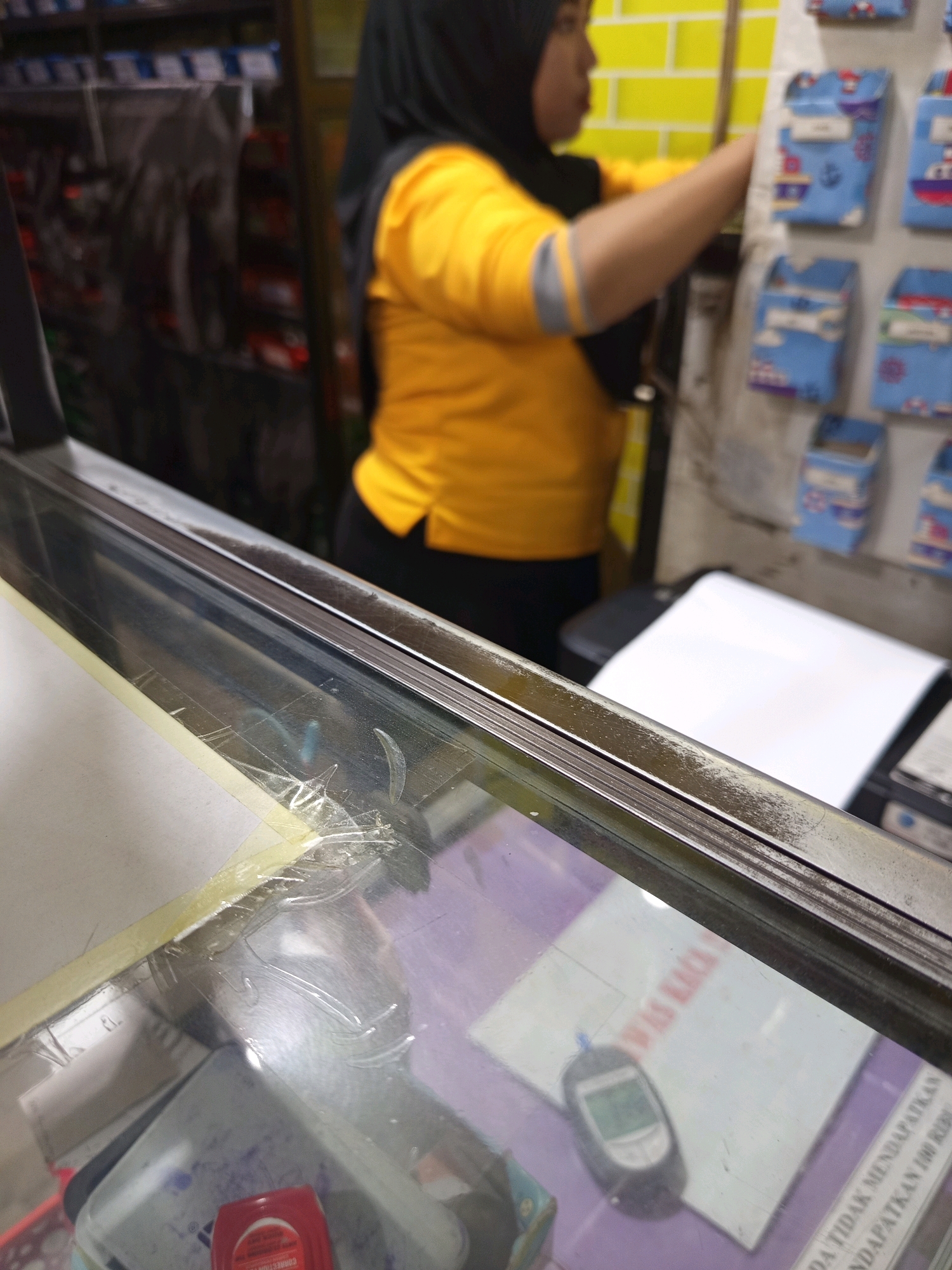 A woman wearing a yellow shirt and a black hijab is arranging small blue fabric pouches on a display board. The pouches appear to be decorated with nautical themes like anchors and boats. The background shows shelves with various items, and a yellow tiled wall. In the foreground, there is a glass counter with some items on it, including a glucometer and some papers. The scene appears to be a shop or a market stall in Muktisari, Indonesia.