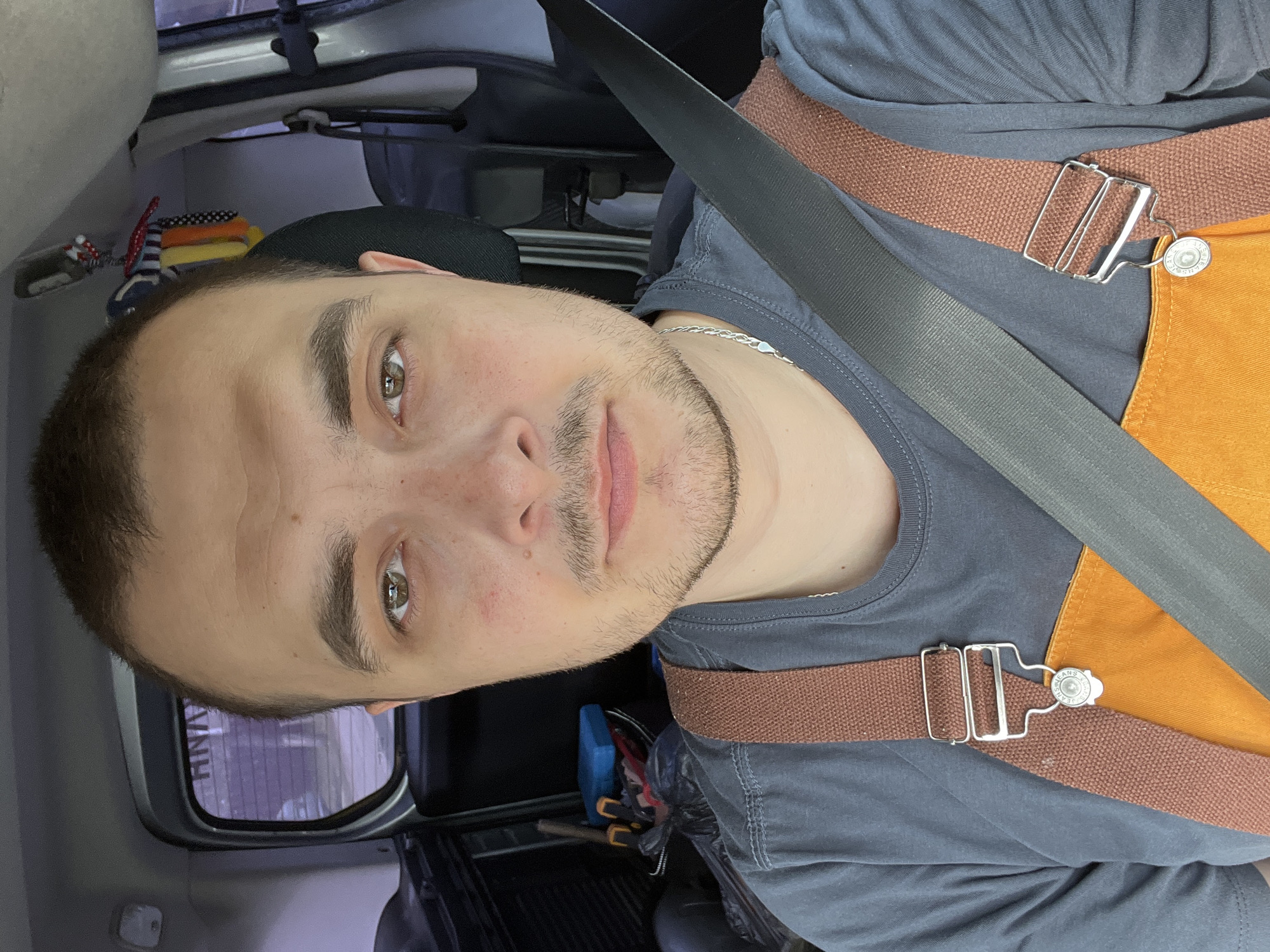 The image shows a young man positioned diagonally across the frame, his head towards the upper left, appearing to take a selfie or looking into the camera while seated in a vehicle. He has short dark hair, light stubble on his chin and upper lip, and light brown or hazel eyes looking directly forward. He is wearing a dark grey t-shirt and a silver chain necklace. Over his t-shirt, he wears an orange garment, possibly an apron or overalls, secured by a brown fabric strap with a rectangular metal buckle. A black car seatbelt crosses his chest.
The background reveals the interior of a vehicle, likely a car or van. To the upper left, behind his head, a stack of colorful folded items (red, white, blue, yellow, orange) is visible. Towards the lower right, behind his shoulder, a blue toolbox and various tools, including pliers and wrenches, are visible on the floor, alongside a black plastic bag. Through a small back window, faint reflections or patterns of a building exterior with vertical lines can be seen, along with some indistinct text that appears to include "ANH". On the silver button of the brown strap, the word "JEANS" is visible, surrounded by small stars. The man has a neutral to slightly serious expression. The lighting suggests it is daytime.