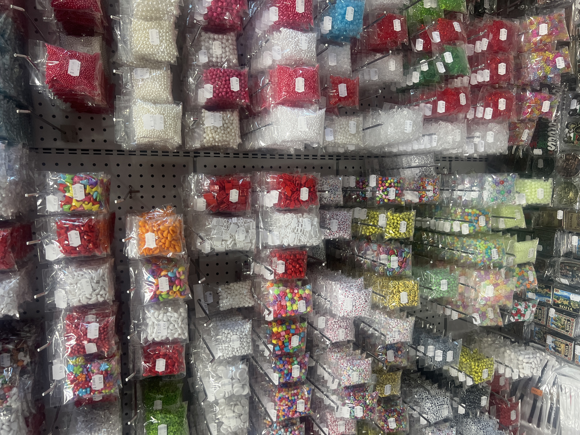 The image displays a retail display of various craft supplies, seemingly beads, in small, clear plastic bags. They are neatly organized on a gray pegboard rack, with multiple rows and columns. The location is Burgas, Bulgaria, as indicated by the 'SOFIA' brand tags visible on the right side of the frame. The beads vary in color, shape, and material, including red, white, blue, green, orange, yellow, and multicolored options. Price tags, labeled in Euros (€), are attached to the front of many of the bags.