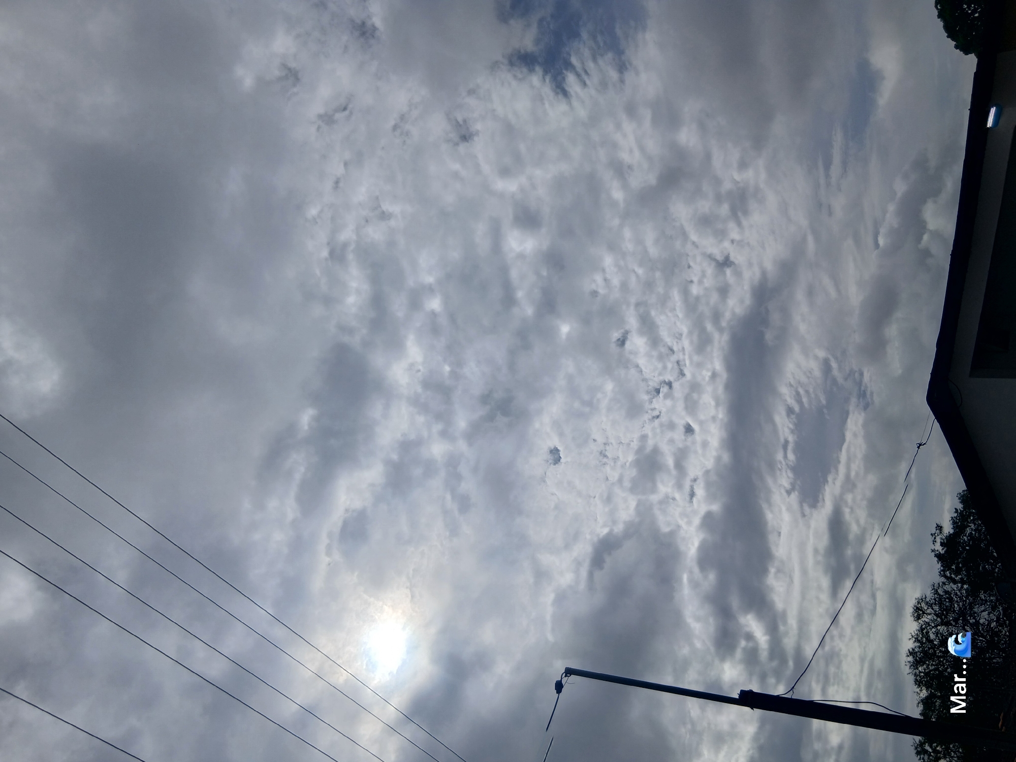 The image depicts a cloudy sky over Juan Griego, Venezuela. The sun is partially visible through a break in the clouds, casting a bright light. Power lines are strung across the upper portion of the frame, running diagonally from left to right. A streetlamp is visible on the left side of the image, its pole extending upwards. In the lower portion of the image, the dark silhouette of trees and the roofline of a building can be seen. Text superimposed on the image reads "Mar..." followed by a wave emoji. The overall atmosphere is overcast, suggesting a day with diffused sunlight.