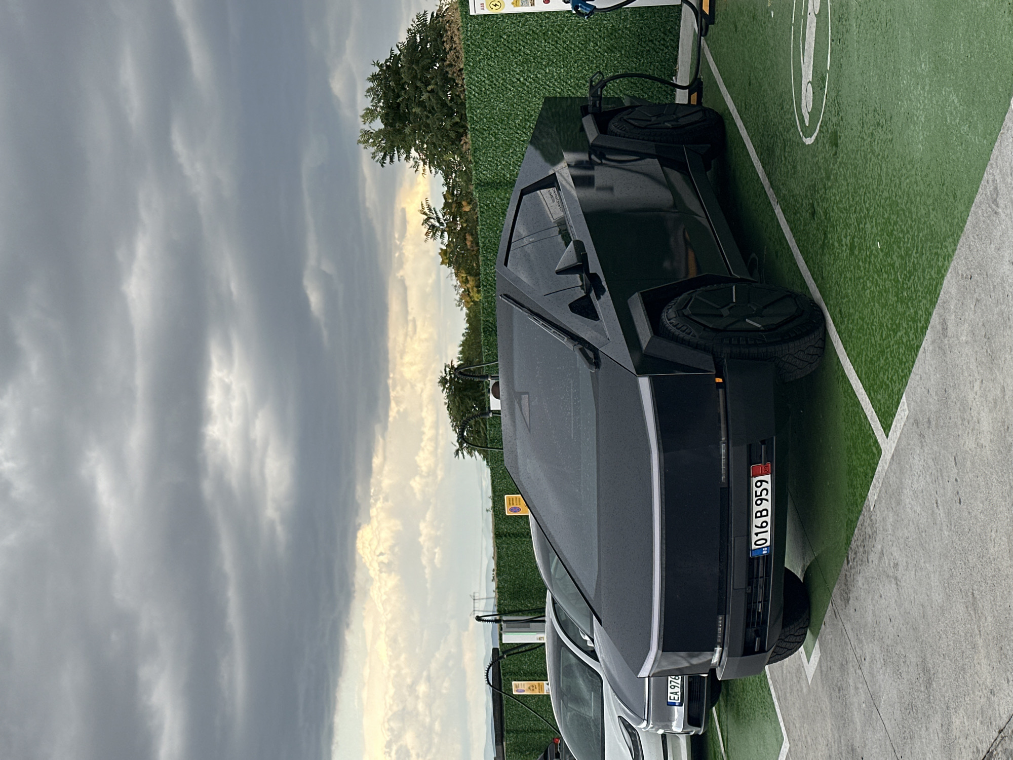 In Polsko Padarevo, Bulgaria, under a cloudy sky, a black Tesla Cybertruck is parked at a charging station. The vehicle is positioned in a marked parking spot with green ground cover, indicating it is an electric vehicle charging area. A charging cable is connected to the Cybertruck from a charging unit mounted on a green hedge. In the background, another electric vehicle, a white Tesla, is also parked at a charging station. The surrounding environment includes trees and a low fence. The license plate on the Cybertruck is visible, displaying "BG 016 B 959". The scene appears to be a public charging facility.