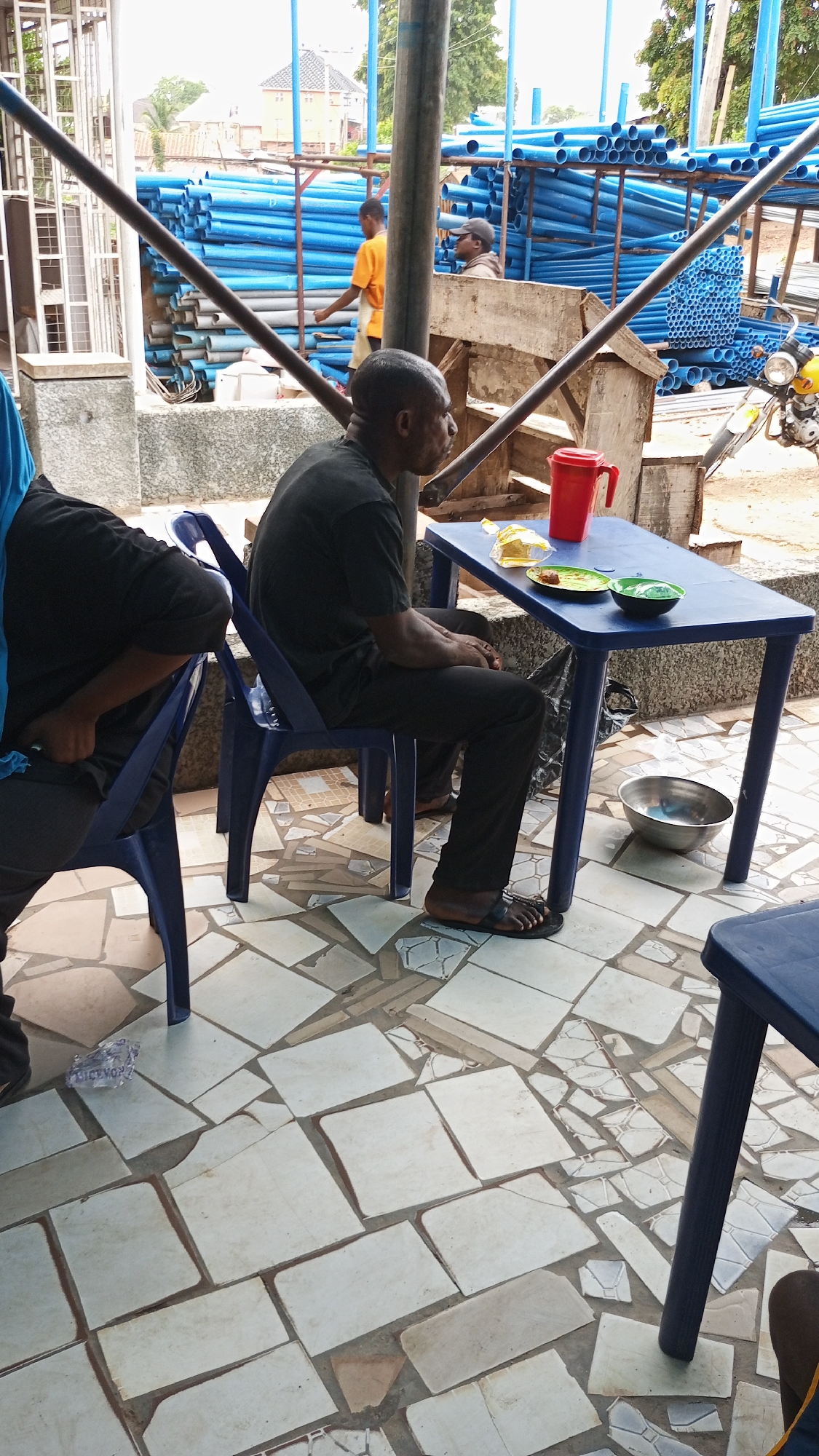 The image shows a man sitting at a blue plastic table, seemingly eating a meal. He's dressed in dark clothing and wearing sandals. On the table, there are plates of food, a red pitcher, and some plastic packaging. A metal bowl sits on the ground nearby. Another person is partially visible, seated to the left.
In the background, there's a stack of blue pipes, suggesting a construction or plumbing supply area. Two other men are visible near the pipes. A building with a brown roof and foliage are in the distance. The flooring is made of broken, mismatched tiles.
Given the location of Lokoja, Nigeria, the scene appears to be an informal eatery or gathering spot near a commercial area, with the blue pipes indicating a business dealing in plumbing or construction supplies nearby. The state of the flooring and the plastic furniture suggest a simple, practical setting.