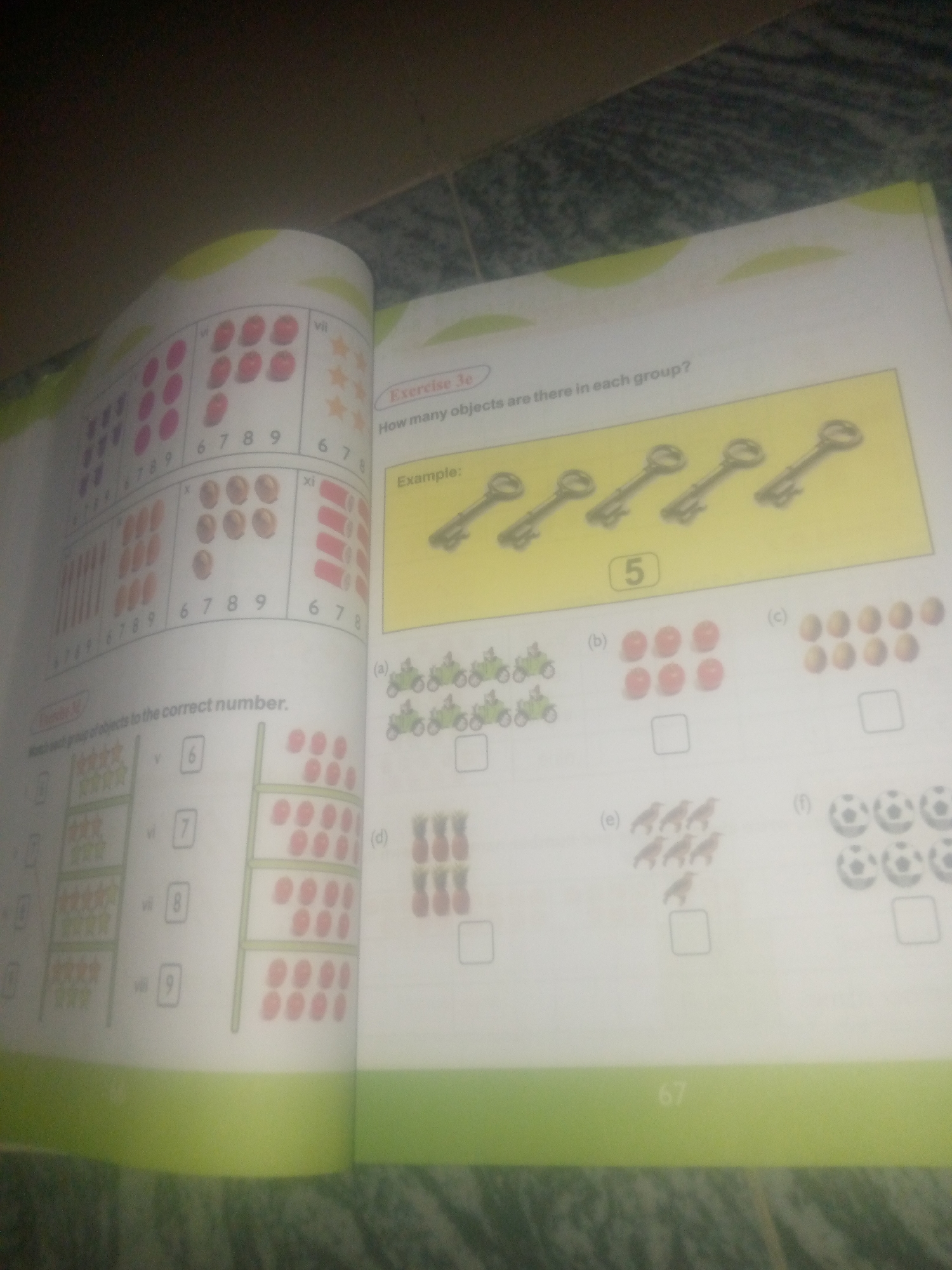 The image shows an open book with two pages visible. The right page features an exercise labeled "Exercise 3e" with the question "How many objects are there in each group?". Below this, there is an example showing five keys in a row with the number "5" in a box. The rest of the right page contains several lettered groups of objects (a, b, c, d, e, f) each with an empty box below it, presumably for the user to write the count.
The left page has an exercise labeled "Exercise IV" titled "Match each group of objects to the correct number." This page displays several rows of objects on the left and numbered boxes on the right. The objects include stars, apples, and what appear to be cylindrical objects. The numbered boxes show the numbers 6, 7, 8, and 9. There is also a vertical arrangement of apples on the far right.
The image is a close-up shot of the book lying on a surface that appears to be a patterned floor or tabletop. The lighting suggests it is an indoor setting. There are no people visible in the image. The image does not contain any information that could be used to determine its location in Jauro Yinu, Nigeria.
