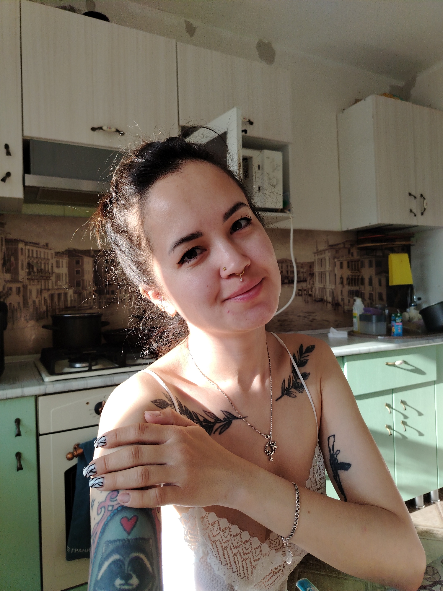 A woman sits in a kitchen. She has dark hair pulled up in a bun, a nose piercing, and tattoos on her arms and chest. She is wearing a white tank top and a necklace. Her left arm is across her chest. The kitchen has light-colored cabinets, a gas stove, and green lower cabinets. A wallpaper with a cityscape design is on the wall behind the stove. There are cabinets above the stove. The location is most likely Noviy, Russia based on a tattoo on the woman's arm that says "За Грань" which translates to "Beyond the Edge".