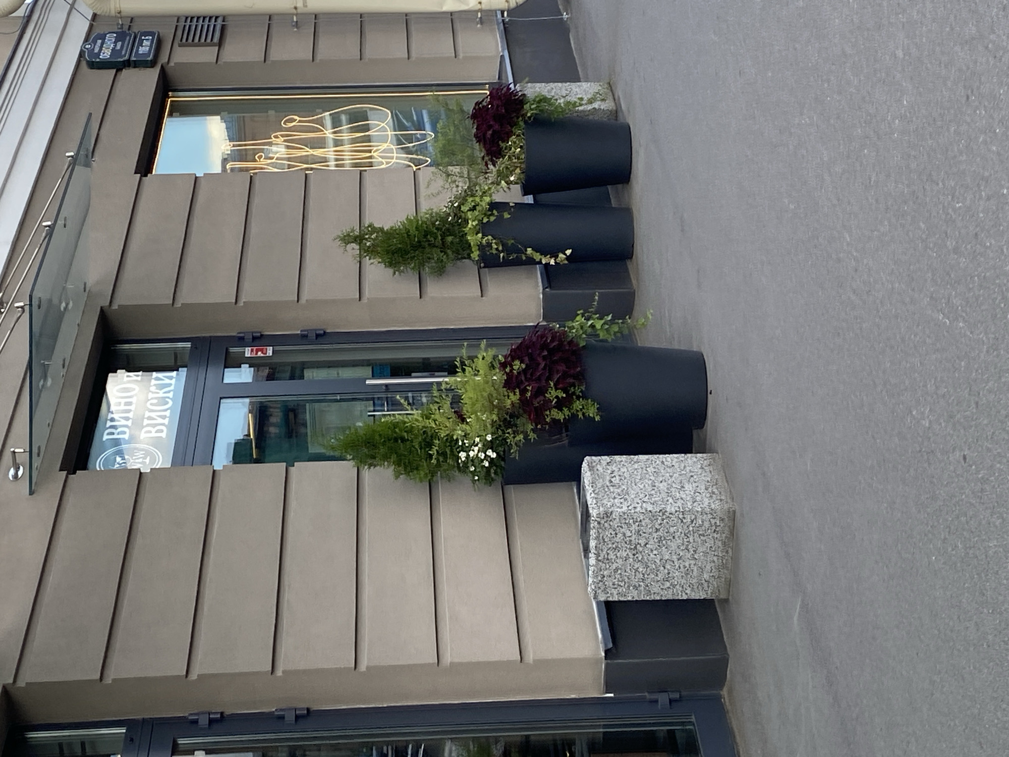 The image shows a storefront on a city street in Saint Petersburg, Russia. The building has a modern design with horizontal beige cladding and large glass windows. A sign above the entrance reads "Вино и Виски" (Wine and Whiskey). In front of the store are several large black planters containing evergreen shrubs and dark red foliage plants, as well as a speckled grey concrete planter. A street sign visible on the building reads "НАБЕРЕЖНАЯ ОБВОДНОГО КАНАЛА" (Embankment of the Obvodny Canal) and "116 Лит. 5", indicating the location. The street appears to be paved with asphalt, and a sidewalk runs along the storefront. The lighting suggests it is daytime, and the sky reflected in the windows is blue. There are no people visible in the frame.