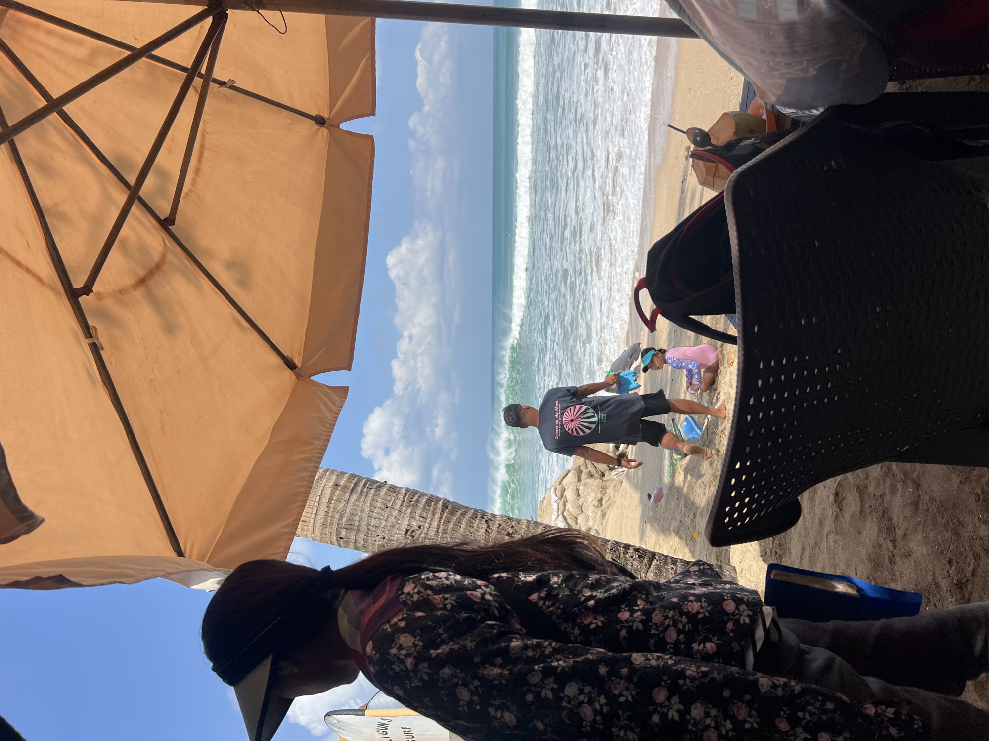 The image depicts a beach scene in Kuta, Indonesia. In the foreground, partially obscured by a large orange umbrella and a black plastic chair, is the back of a woman wearing a floral print long-sleeved shirt and a visor. To her right, a man in a dark t-shirt and shorts stands facing the ocean, holding a blue plastic shovel and bucket. A young child wearing a pink swimsuit and a blue visor is seated on the sand near him. The ocean features moderate waves, with some surfers visible in the distance. A palm tree stands to the left of the man. In the middle ground, there are sandbags and a surfboard. A coconut with a straw and spoon rests on a table in the lower right corner of the frame. The overall environment suggests a relaxed beach day under a bright, partly cloudy sky.