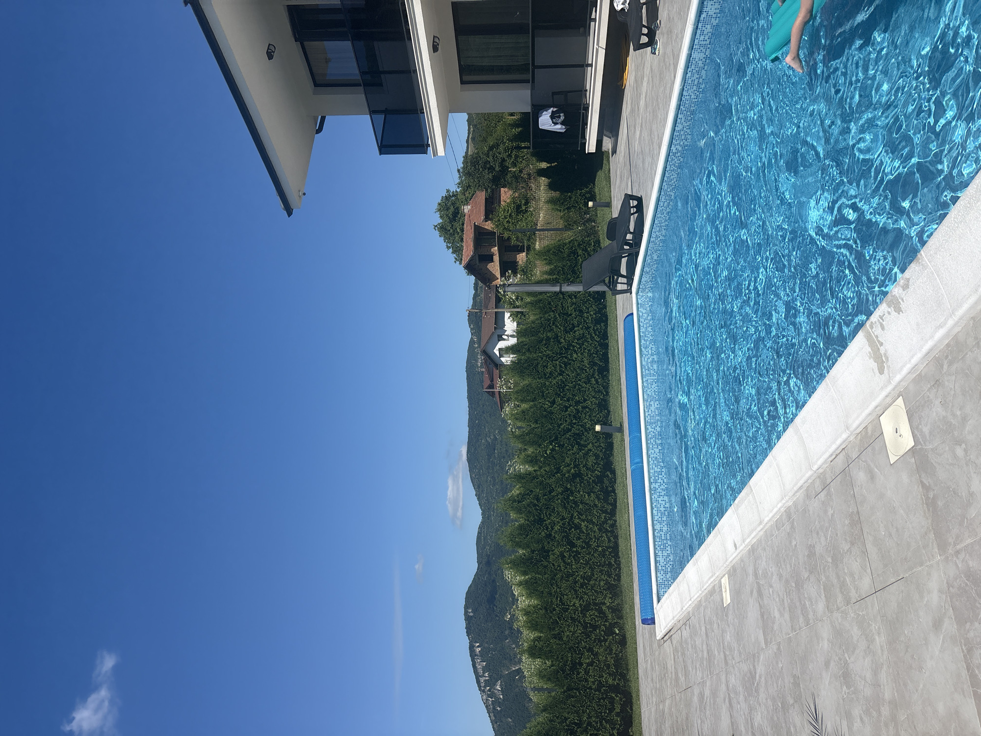An outdoor swimming pool is shown under a clear blue sky with a few wispy clouds. The pool has a blue tiled liner and a white coping. A blue pool cover is partially visible on the left side of the pool. The pool is surrounded by a gray tiled patio. Two black lounge chairs are placed on the patio next to the pool. A person's legs and a portion of a green inflatable float are visible in the water on the right side of the pool. In the background, a row of tall green cypress trees lines the edge of the property. Beyond the trees, there are rolling green hills and mountains, with several houses and buildings scattered among them. A modern white house with a black-trimmed roof and balconies is situated on the right side of the image, overlooking the pool and surrounding landscape. The overall scene depicts a sunny day in a residential area with a view of natural scenery.