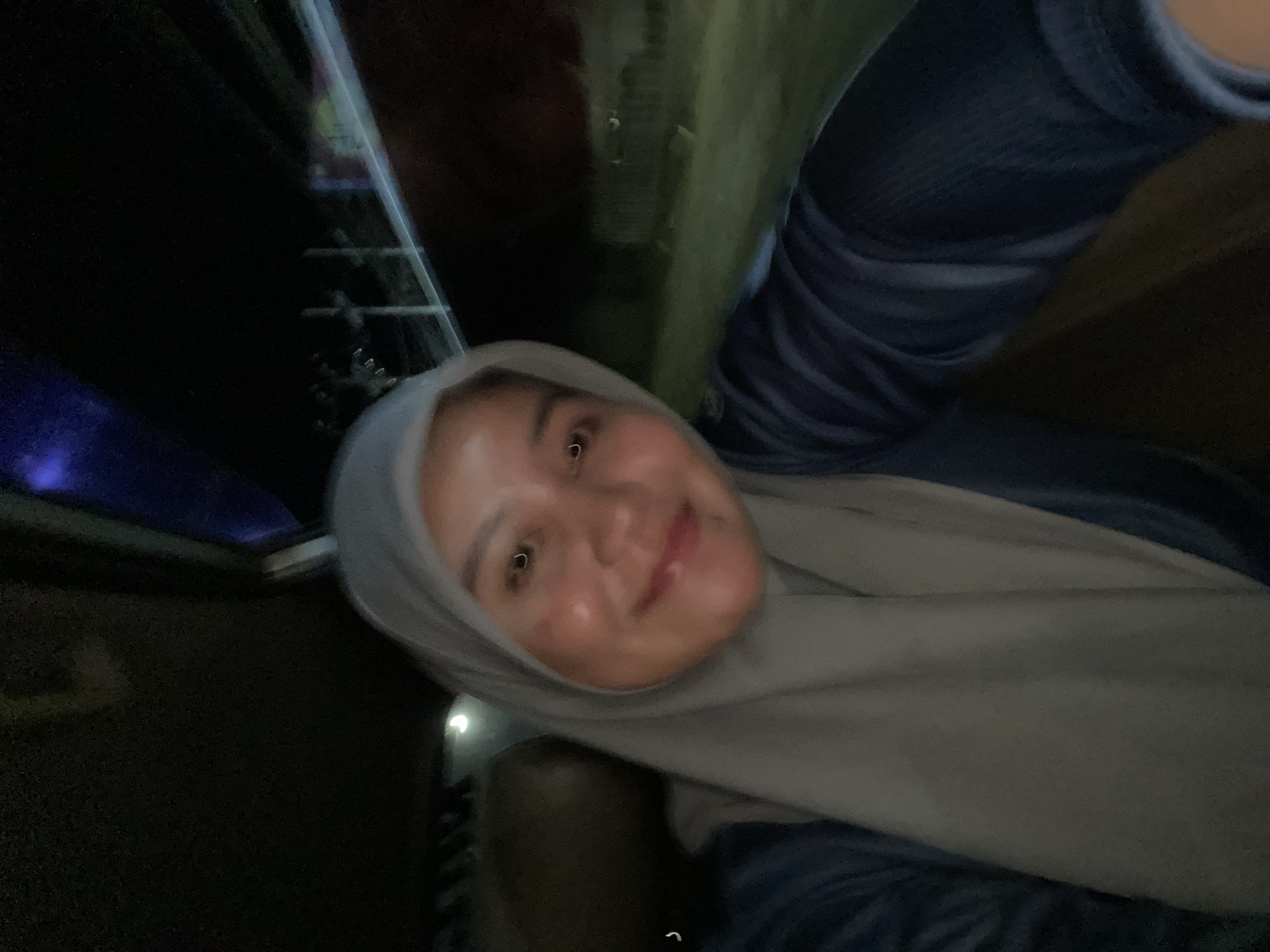 The image is a close-up, vertically oriented selfie of a young woman wearing a grey hijab and a dark blue shirt. Her face is the primary subject, with her head tilted and a slight smile on her face. The background is dark and indistinct, with hints of blurry lights and structures, suggesting an indoor or nighttime outdoor setting. The lighting is somewhat dim, with a slight sheen on her skin, possibly from makeup or ambient light. One of the crops shows a faint hint of what might be a sign or poster with colorful elements in the dark background. The overall mood appears casual and relaxed. The image was taken in Sungai Paring, Indonesia.