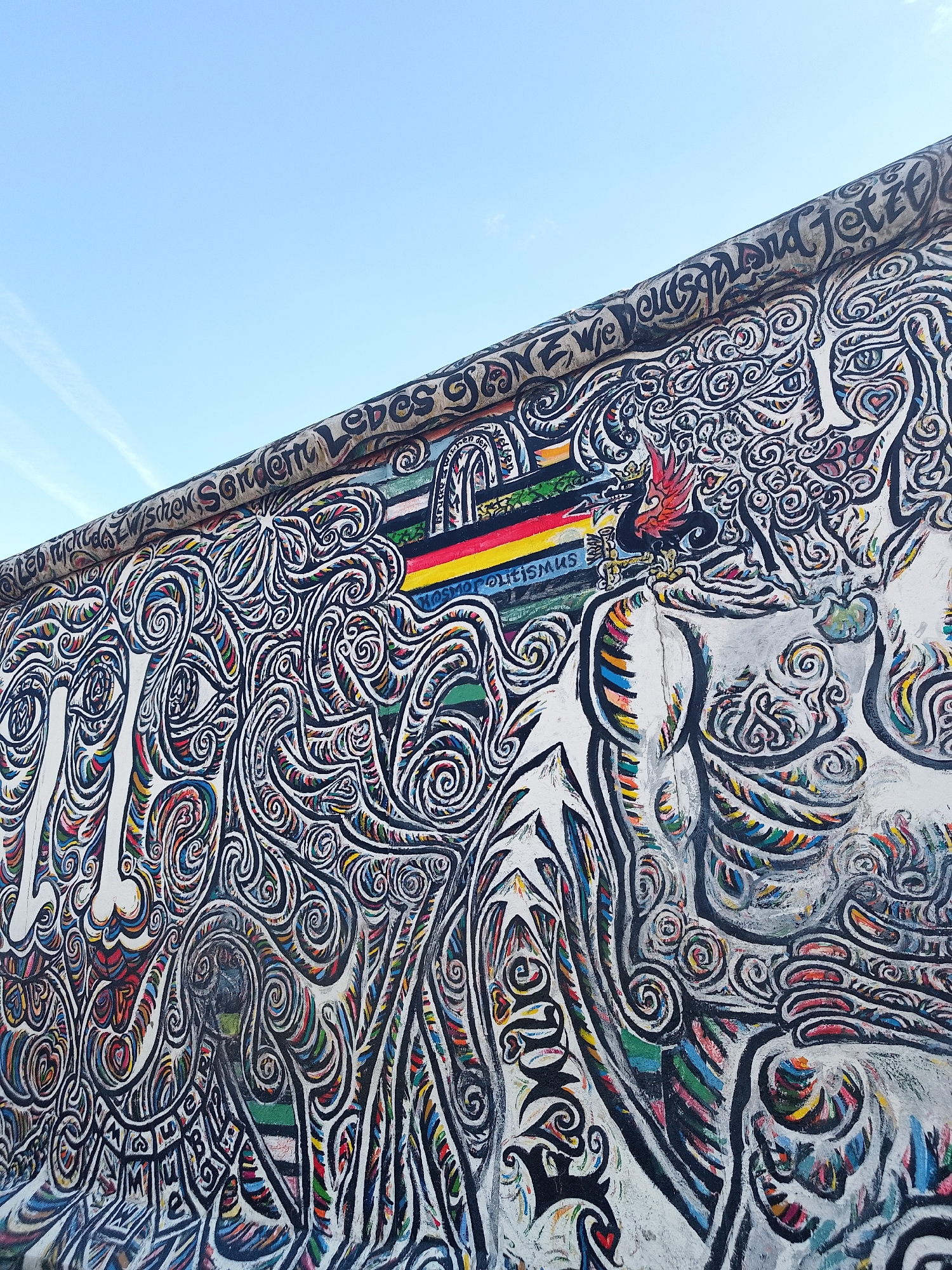 The image shows a section of the Berlin Wall, located in an unknown city and country. The wall is covered in graffiti and artwork, featuring swirling patterns and various shapes outlined in black. A horizontal band of color in the German flag colors of black, red, and gold, along with the word "Kosmopolitismus" are visible. In the upper portion of the wall is lettering in German. The sky, visible in the upper left, is blue.