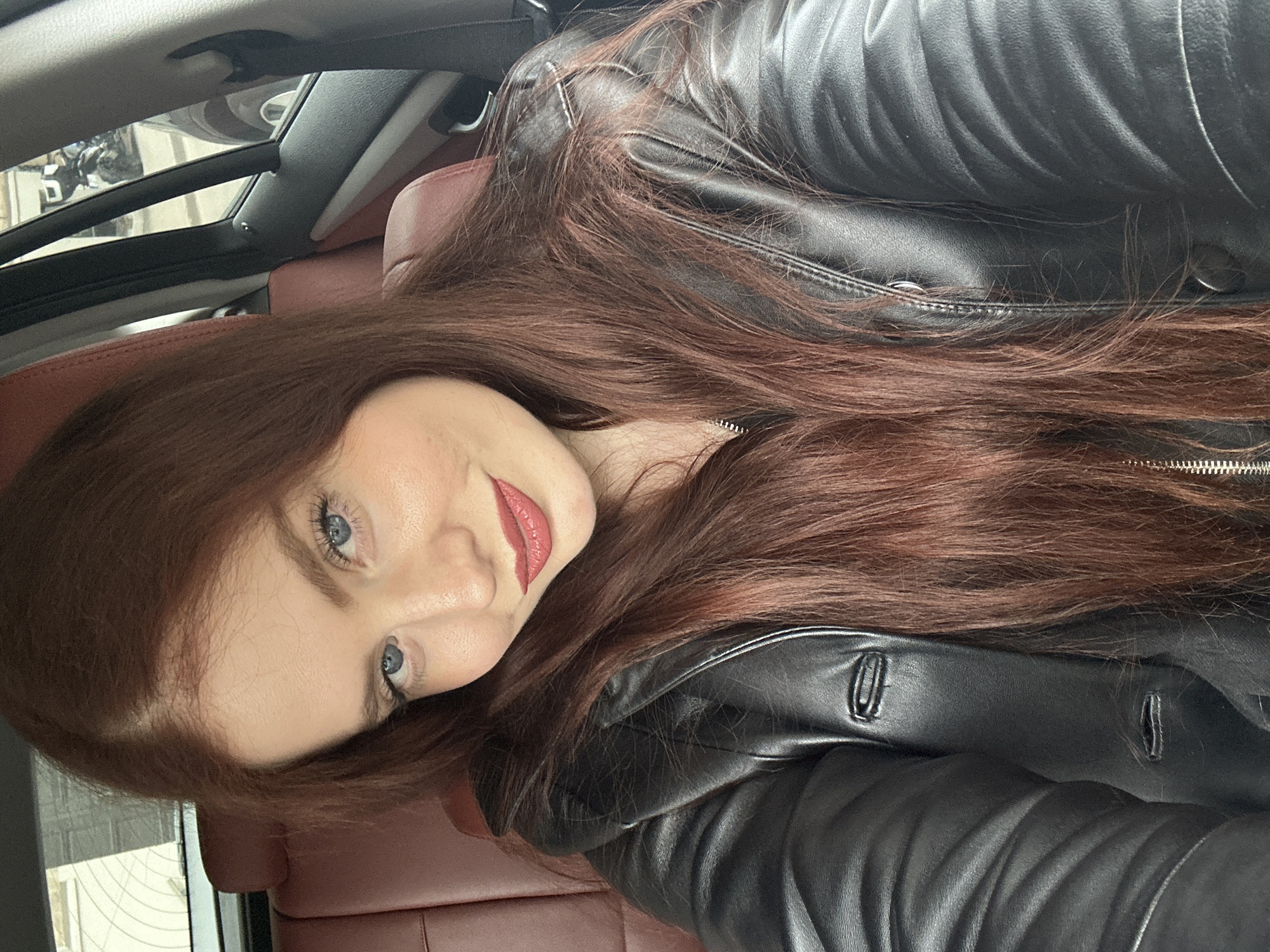 A young woman with light skin, red lipstick, and blue eyes, is inside a car. She has long, auburn hair, and is wearing a black leather jacket with a zipper. The car has red leather seats and a sunroof. The image was taken in Riga, Latvia. The exact location in Riga cannot be determined from the provided image.