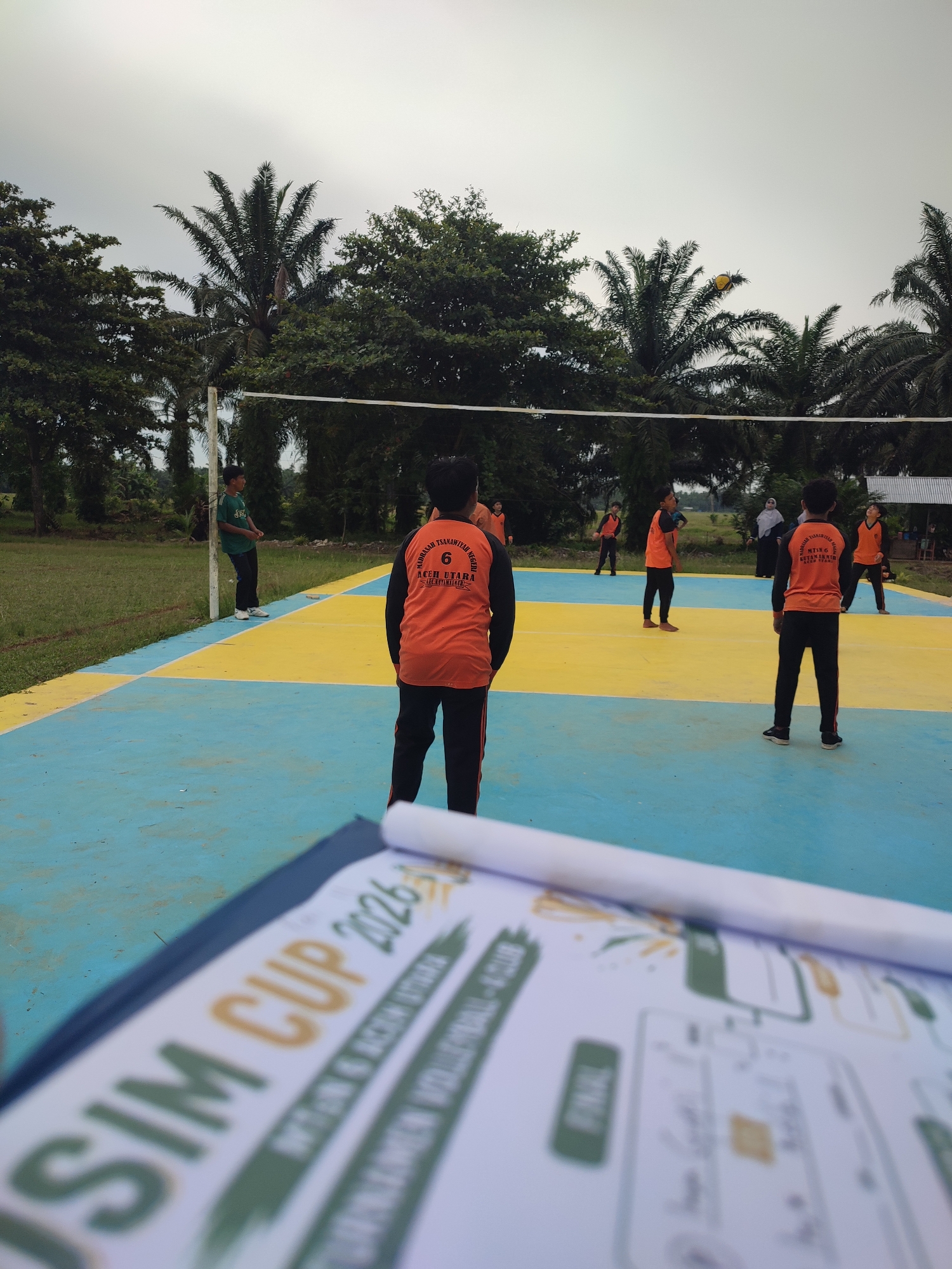 The image depicts an outdoor volleyball game in progress on a court with blue and yellow markings, located in Blangriek, Indonesia. Several young males, dressed in athletic attire, are actively participating in the game. Some are positioned on the court, seemingly preparing to receive or serve the ball, while others are in the background, observing or waiting for their turn. A yellow volleyball is visible in the air near a palm tree. The environment is characterized by lush green trees, including prominent palm trees, and a cloudy sky overhead. In the foreground, a clipboard with a document is partially visible, suggesting that the game might be part of an organized event. The overall scene conveys a sense of youth sporting activity in a tropical setting.