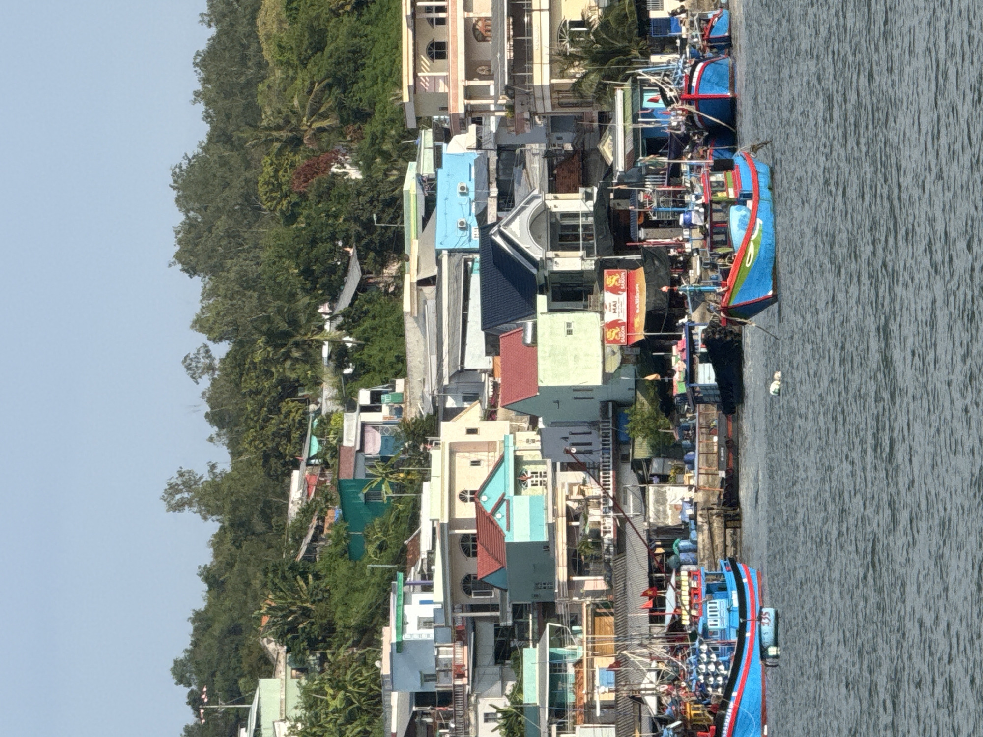 The image depicts a coastal scene in Nha Trang Ward, Vietnam, featuring a densely packed hillside of houses and buildings overlooking a body of water. The architecture is varied, with many houses painted in bright, diverse colors like teal, light blue, and cream, often featuring multiple stories and balconies. Some buildings have red tiled roofs, while others have flat, modern designs. Greenery, including trees and lush vegetation, covers the hillside, partially obscuring the structures.
In the foreground, the water is choppy with ripples. Several fishing boats are visible, most notably a bright blue and red boat in the center of the frame, and another larger blue and red boat further to the right. These boats appear to be docked or anchored. The boats are equipped with numerous fishing nets and equipment, suggesting an active fishing community. There are no people clearly visible in the water or on the boats. In the background, the hillside is densely populated with houses of various sizes and colors, some appearing weathered and others more modern. A clear blue sky dominates the upper portion of the image. The overall impression is of a vibrant, lived-in coastal community.