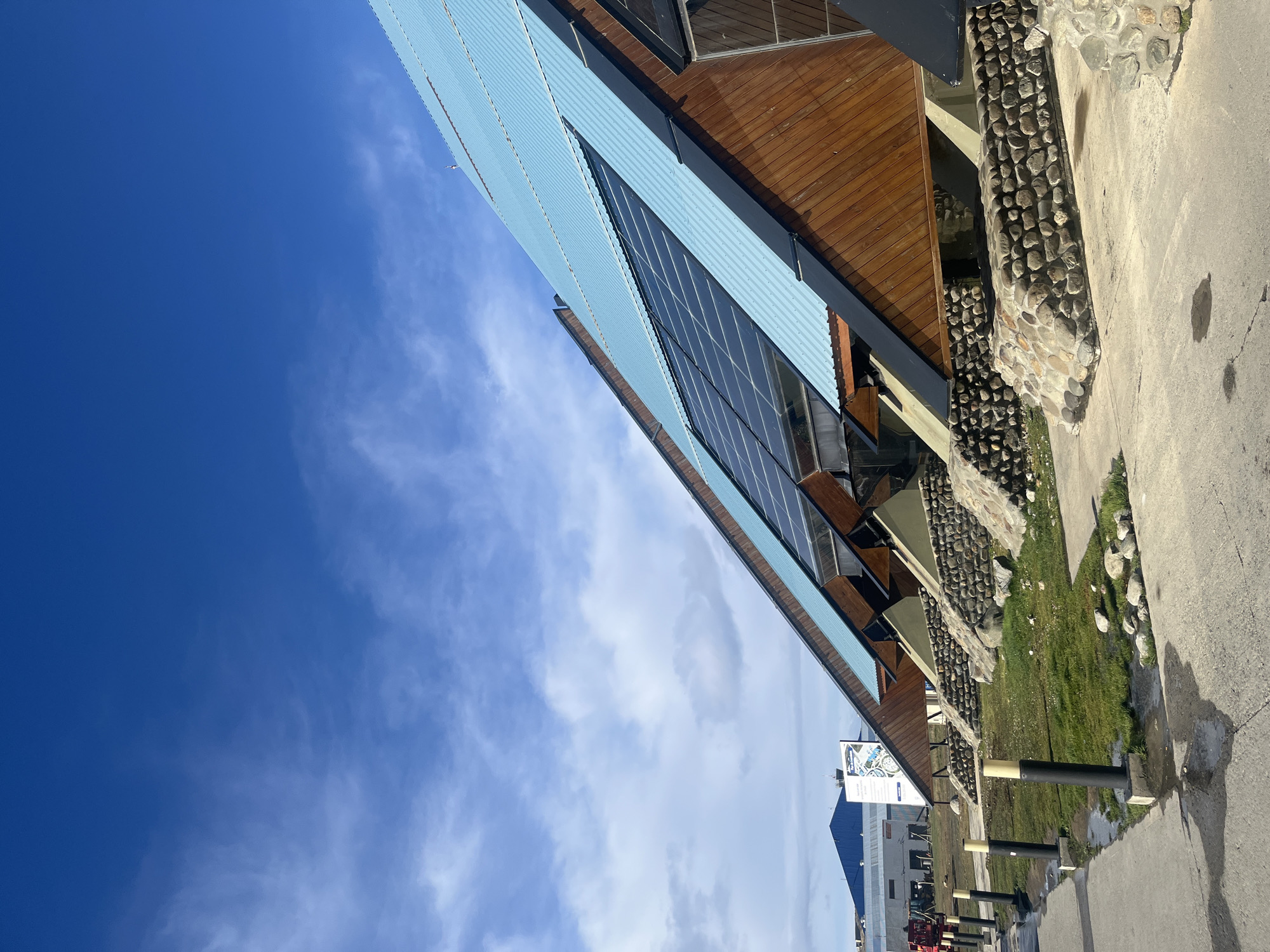 The image depicts an upward-angled view of a building in Ushuaia, Argentina, beneath a sky ranging from deep blue to cloudy white. The building features a light blue corrugated metal roof, brown wooden paneling, and a series of windows. Stone and grass are visible along the side of the building, extending to a concrete walkway with black posts. Distant buildings and infrastructure are seen in the background.