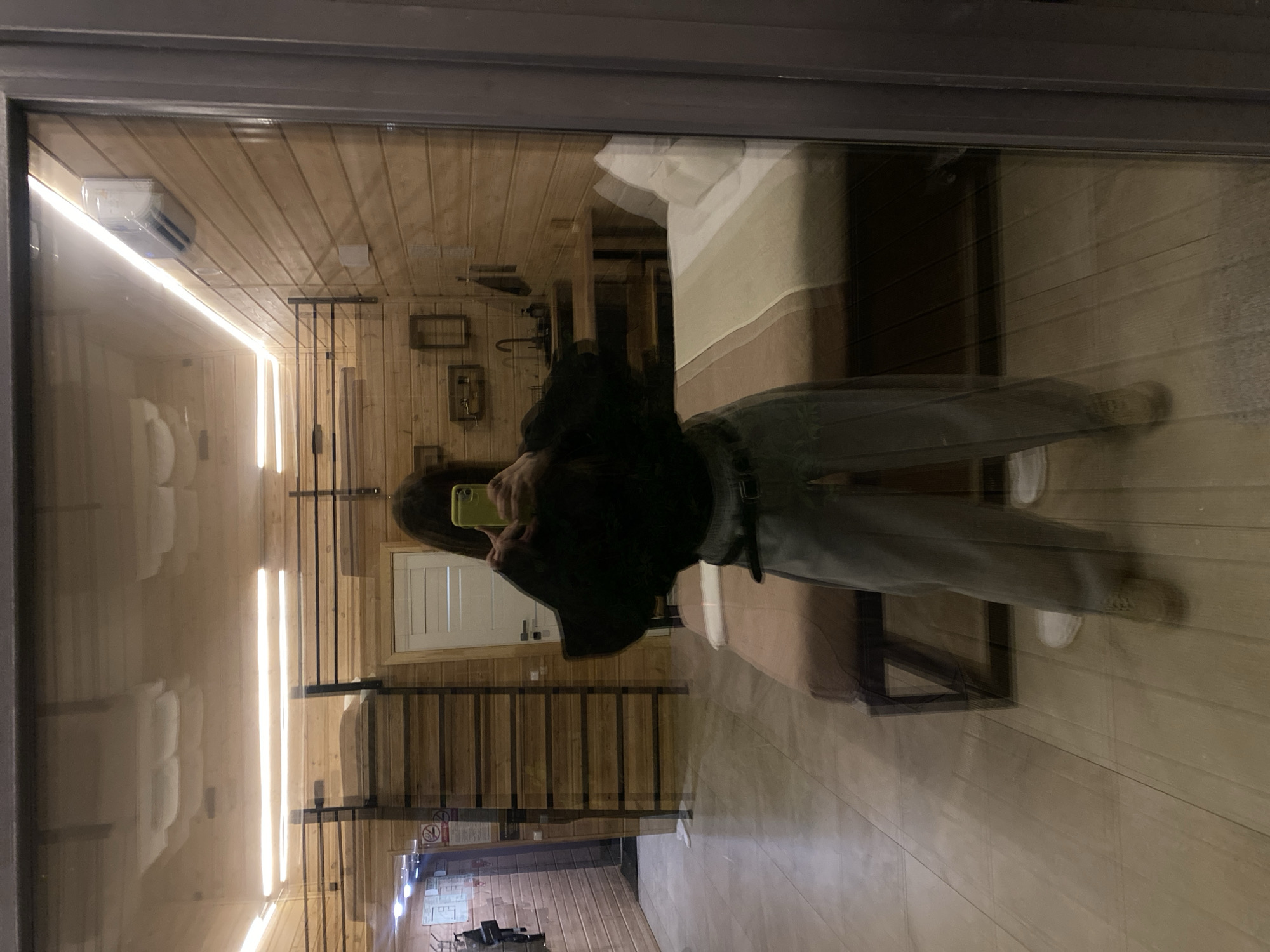 A person is reflected in a large mirror or glass panel, centered within a modern, wood-paneled room. The individual, whose face is obscured by dark hair and a light green smartphone held up, is taking a mirror selfie. They are wearing a black long-sleeved top, light-colored wide-leg pants secured with a black belt, and beige low-top shoes.
The room features light wood paneling on the walls and ceiling, with a light-tiled floor. Overhead, several rectangular flush-mount lights are active, alongside an LED strip light running along the right wall. On the left side of the reflection, a dark, multi-rung ladder-like structure is visible against the wall, next to a white door. A "No Smoking" sign is partially visible on the far left wall. On the right side, a bed with white pillows and a striped light grey and beige duvet cover is present. Above the bed on the right wall, an air conditioning unit is mounted. Further back and to the right, a small kitchenette area with a dark countertop, a sink with a black faucet, and shelves with decorative items is discernible. The scene is located in Bakhtiyary, Russia.