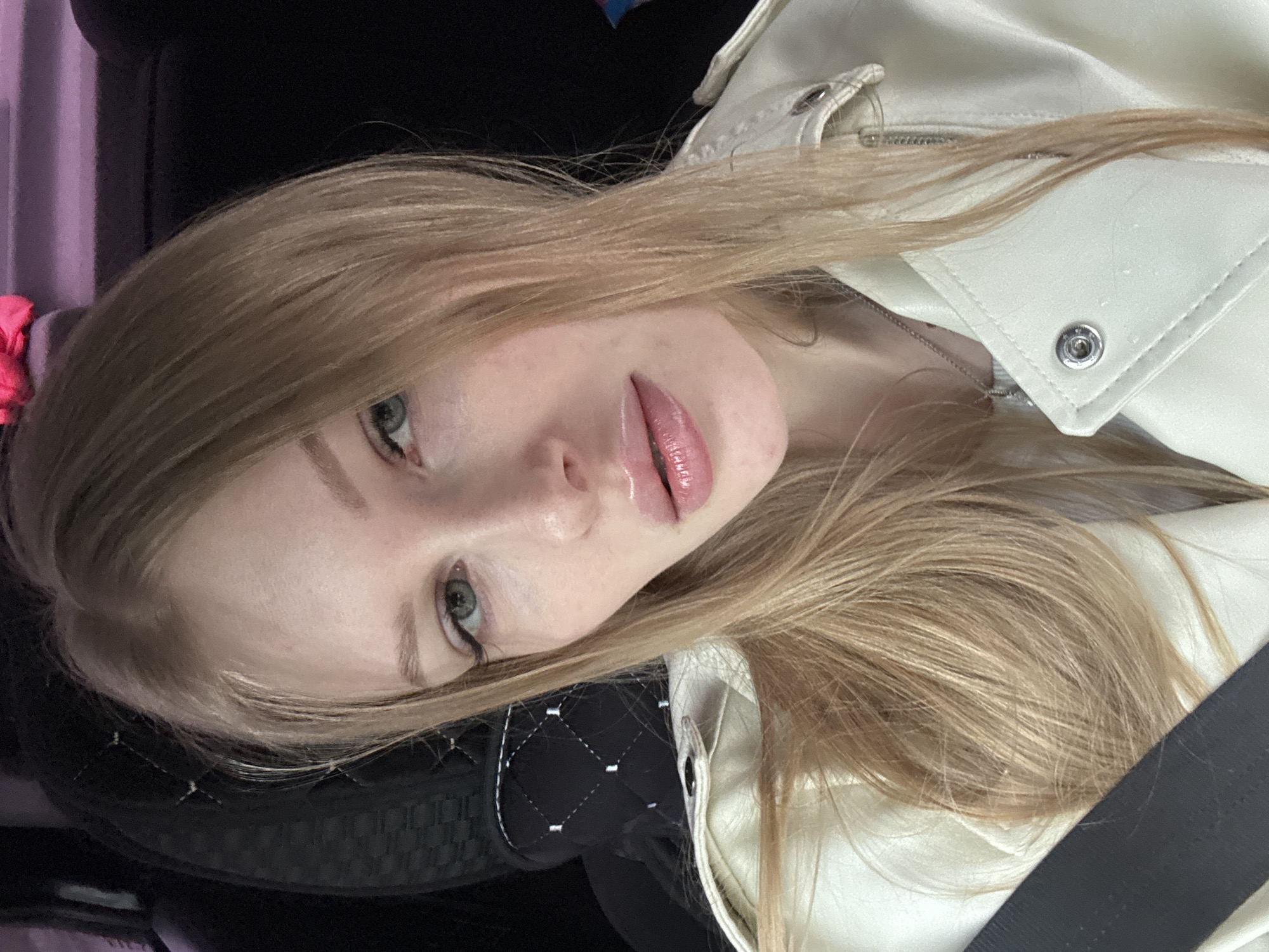 A close-up portrait of a young, light-skinned woman with long, light brown hair and green eyes. She is wearing a white leather jacket and a seatbelt. The background is blurred, suggesting she is in a vehicle. Based on the limited visual information, it's not possible to confirm the location as Perm, Russia.