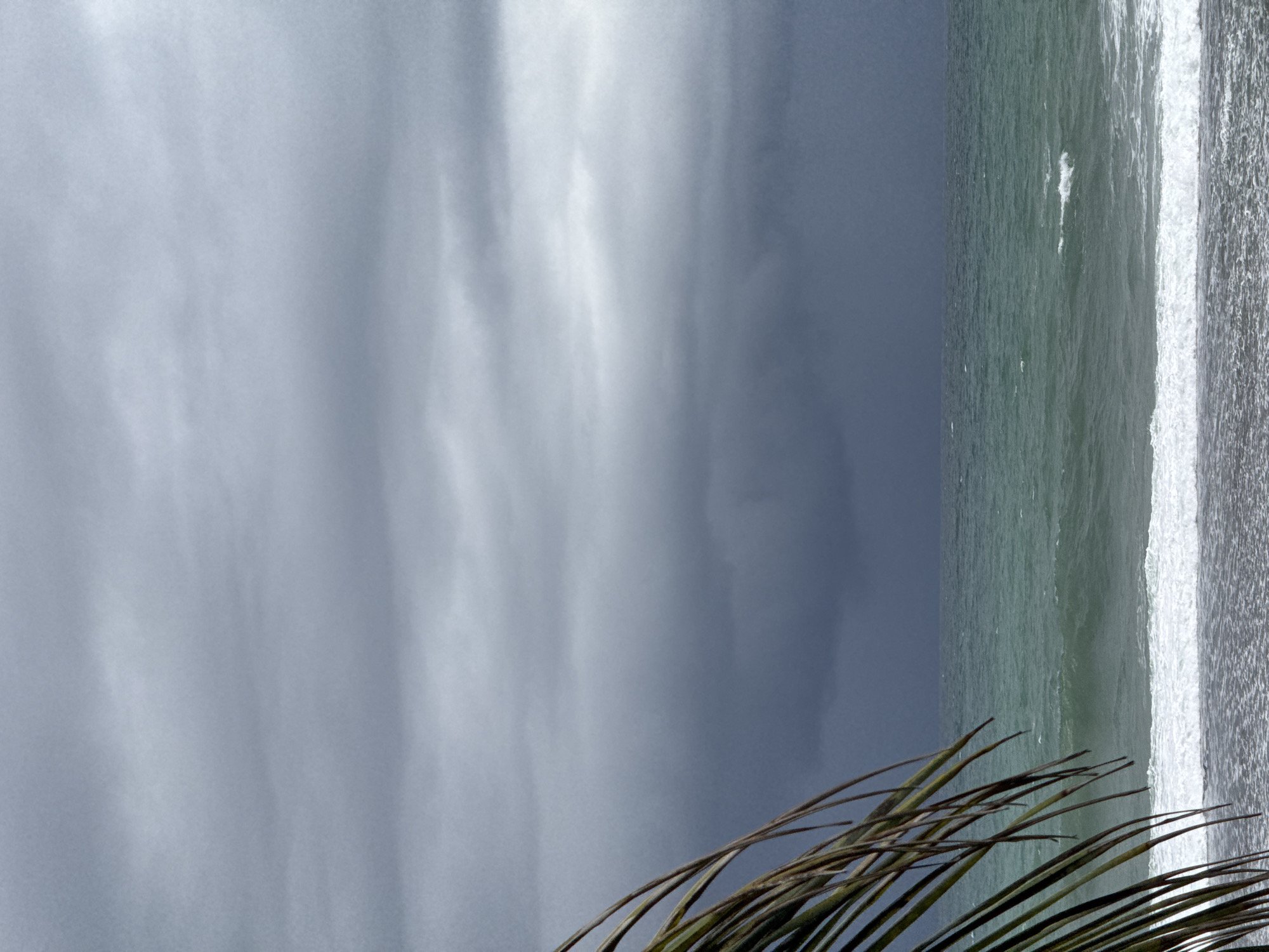 A dark, cloudy sky looms over the choppy, teal-colored ocean near the shore of Ahangama, Sri Lanka. In the foreground, the fronds of a palm tree are visible on the left, partially obscuring the view of the sea and sky. The waves are breaking onto a sandy beach, creating a white line of foam. The overall impression is of an approaching storm.