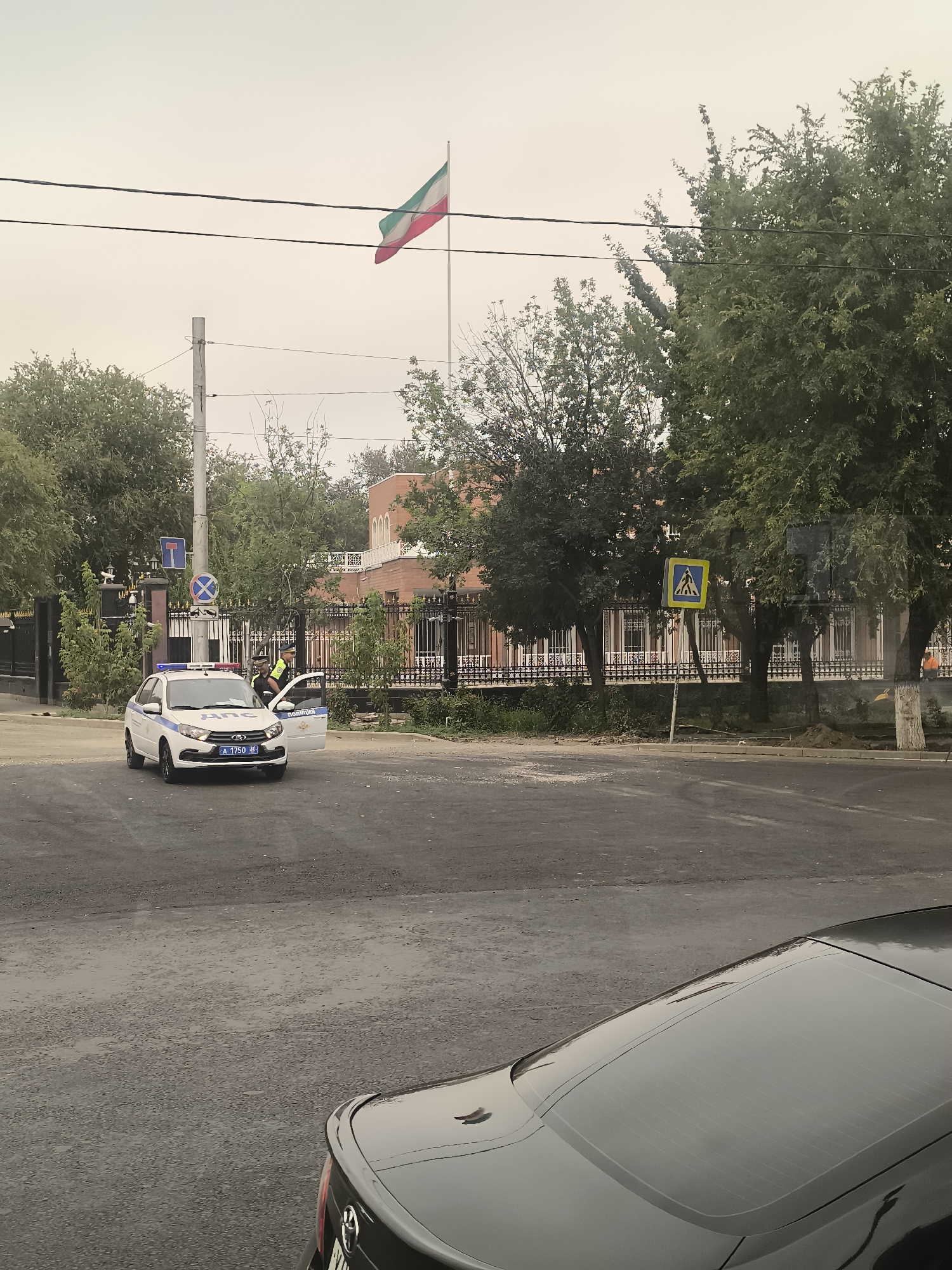 The image depicts a street scene in Astrakhan, Russia, with a police car stopped at an intersection. The police car, a white sedan with blue and red flashing lights, has two officers standing near it. One officer is in the driver's seat, and another is standing by the open passenger door, wearing a yellow vest over a dark uniform. The car has the word "ПОЛИЦИЯ" (POLITSIYA - POLICE) written on its side.
In the background, there is a tall flagpole flying a tricolor flag, which appears to be the flag of Iran. Behind the flagpole and the intersection is a building with an arched entrance, likely an embassy or consulate, with a black metal fence in front of it. A pedestrian crossing sign is visible on the right side of the image. The street appears to be wet, suggesting it may have recently rained. A portion of a dark-colored car is visible in the foreground, partially obscuring the view of the street. The sky is overcast and hazy.