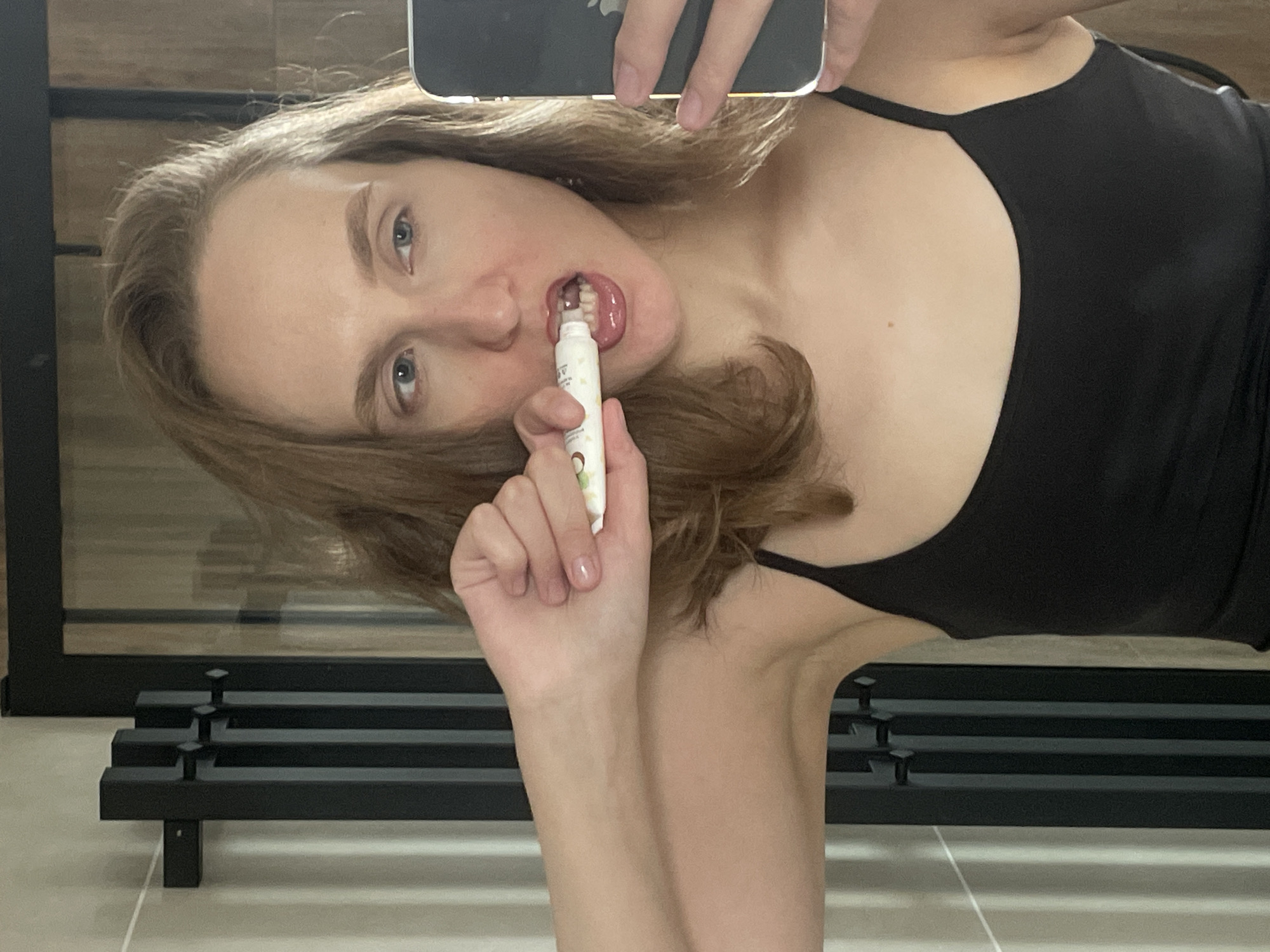 This rotated mirror selfie, taken in an indoor setting, features a woman from the chest up. She has light brown hair and blue-grey eyes, and is wearing a black tank top or sports bra. The image appears to be taken with an iPhone, indicated by the partially visible Apple logo on the phone she is holding. The woman is holding a small, white tube-like object in her mouth, possibly applying a cosmetic product such as lip gloss or teeth whitener. Her right hand, with lightly polished fingernails, is visible holding the tube. The tube has a small green icon, possibly a leaf or coconut, and some illegible text printed on it. In the background, a black metal structure with horizontal bars, possibly a bench or part of a rack, is visible. The floor is light-colored. The overall scene suggests a personal moment, likely in a private space such as a bathroom or changing room in Podshivalovo, Russia. The woman's expression is neutral and focused on the application.