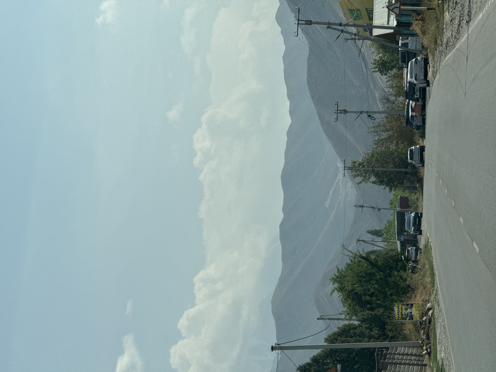 The image depicts a view of a road leading towards a mountainous landscape. The foreground shows a paved road with white lane markings. To the left of the road, there is a wooden fence, some trees, and a yellow sign with Russian text. Several cars are parked along the right side of the road. In the background, a series of rolling hills and mountains are visible, fading into a hazy blue sky with scattered clouds. Utility poles with wires are present along the road, indicating infrastructure in the area. The overall scene suggests a rural or semi-urban setting on the outskirts of Tash-Moinok, Kyrgyzstan. There are no people visible in the image.