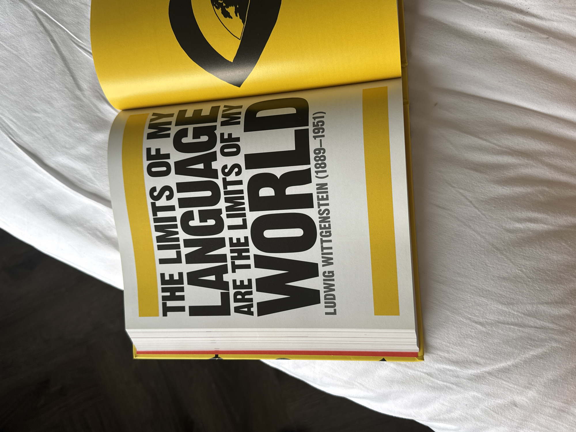 The image shows an open book with a quote by Ludwig Wittgenstein. The quote reads: "THE LIMITS OF MY LANGUAGE ARE THE LIMITS OF MY WORLD." Below the quote is Wittgenstein's name and dates: "LUDWIG WITTGENSTEIN (1889-1951)". The text is in a bold, black sans-serif font against a white background. The pages of the book have a yellow border at the top and bottom. The book is open on a white, rumpled surface that appears to be a bedspread or similar fabric. The background is out of focus and dark on the left, suggesting a dimly lit room. There is a partial view of another page on the right, which is solid yellow with a black graphic element. There are no people or animals in the image. The image does not provide any information about the location of the city of Da Loc, Vietnam.