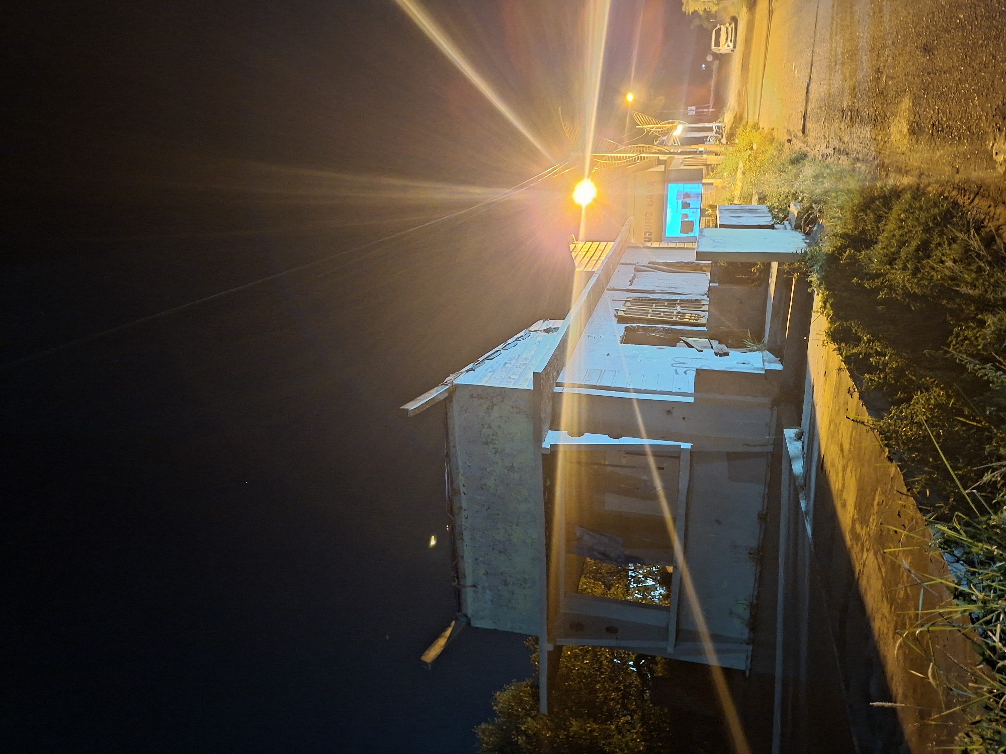 At night, a poorly lit street scene in Lhokseumawe, Indonesia. A white, partially constructed building with exposed framing is positioned on a sidewalk. The building is lit by the glare of a nearby street light, which casts bright beams into the night sky. Grass and foliage grow alongside a cement curb next to the building. Further down the street, another building is visible with the words "WARUNG SAMA" in blue lettering. A vehicle is parked on the side of the street beyond that building. The background sky is dark.