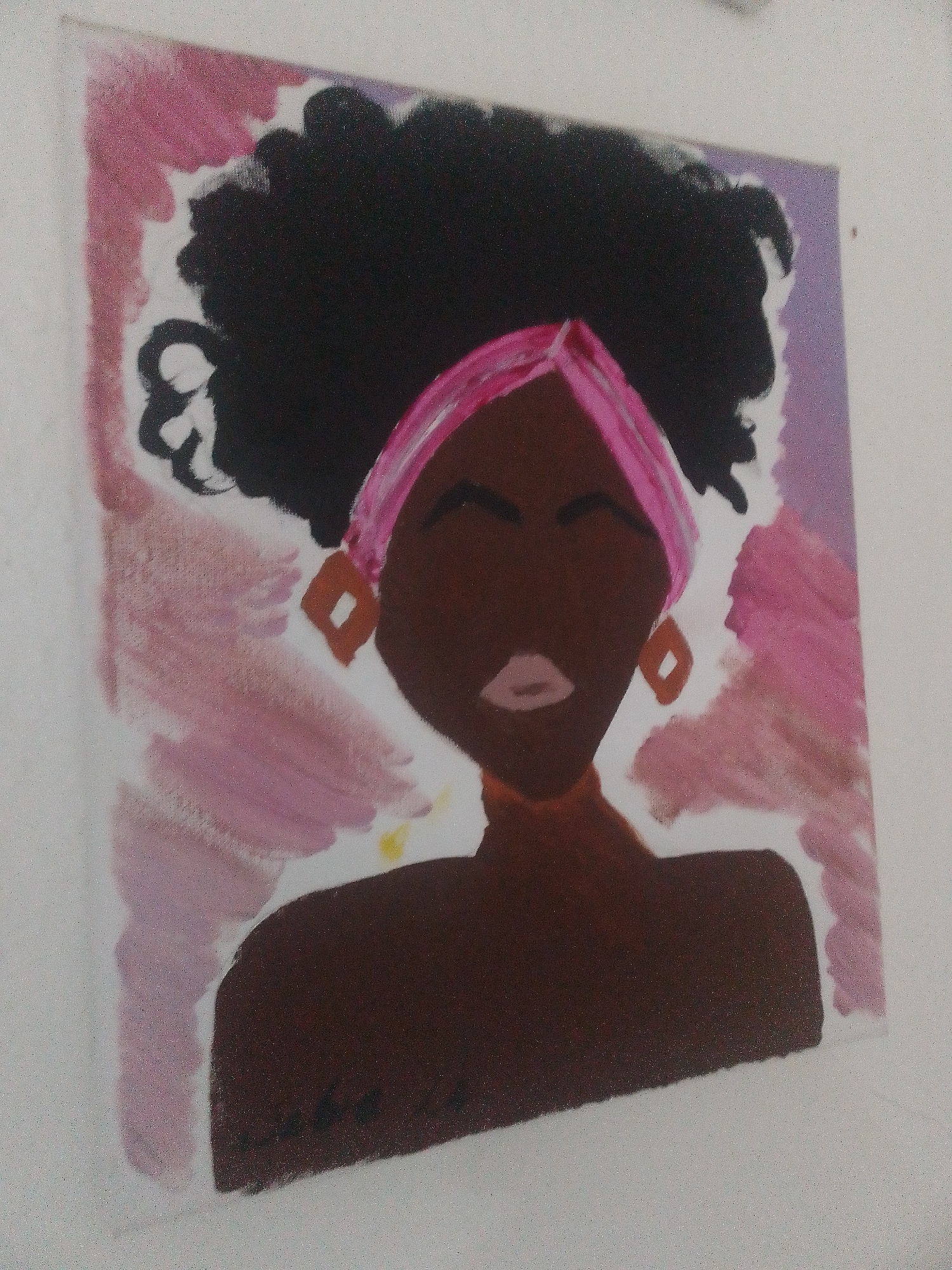 The scene, located in Abuja, Nigeria, captures a framed acrylic painting hanging on a plain white wall. The artwork depicts a stylized front-facing portrait of a person with dark brown skin, large voluminous black Afro-style hair, and a bright pink headwrap. The face features closed eyes represented by thin dark lines, light pink full lips, and rhomboid-shaped orange-brown earrings. A similar colored choker is visible around the neck. The background of the painting is an abstract wash of pink and purple brushstrokes, with white visible around the edges of the hair. The photograph is taken from a slightly low angle, looking upwards and towards the right side of the canvas, revealing a subtle texture on the white wall.