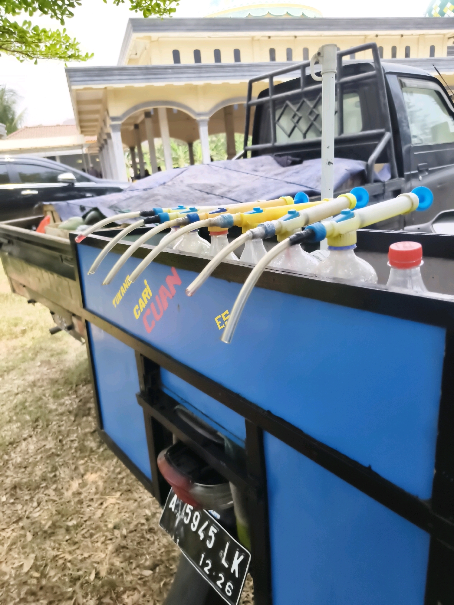 In Puloampel, Indonesia, a pickup truck modified to dispense fuel is parked outdoors. The truck bed contains a blue box-like structure with several bottles filled with liquid, likely fuel, inserted into it. Each bottle has a tube attached, leading to a hand pump mechanism. The text "TUKANG CARI CUAN" is visible on the blue structure. A license plate reading "A 5945 LK 12.26" is attached to the rear of a motorcycle parked behind the truck. In the background, a building with arched windows and a dome, possibly a mosque, is visible. The ground is covered in dry grass and leaves.