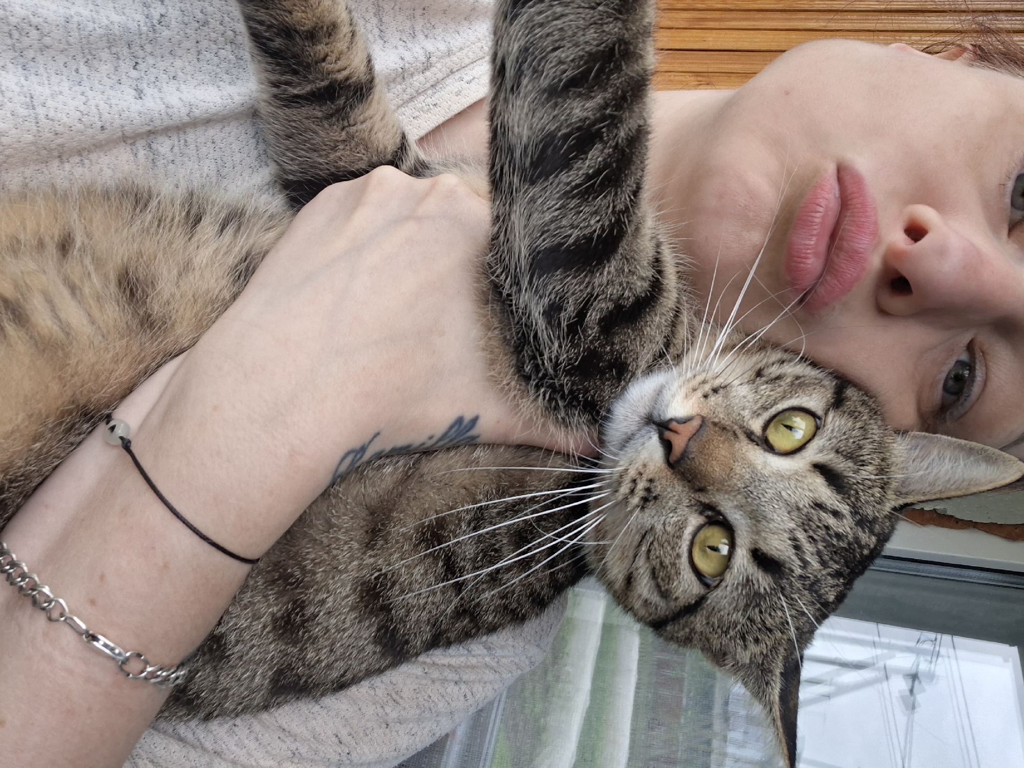 A close-up selfie shows a person holding a tabby cat. The person's face is partially visible, with their lips pursed for a kiss. The cat's head is tilted, with its large yellow eyes looking directly at the camera. The cat's body is wrapped around the person, with its paws and tail visible. The person is wearing a light-colored sweater, and a tattoo is visible on their arm. A black string bracelet and a silver chain bracelet are also visible on the person's wrist. The background appears to be a window with blinds, suggesting an indoor setting with an outdoor view. The image quality is fair, with some digital noise. The setting is likely a residential area within Забалоцкі сельскі Савет, Belarus, given the context.